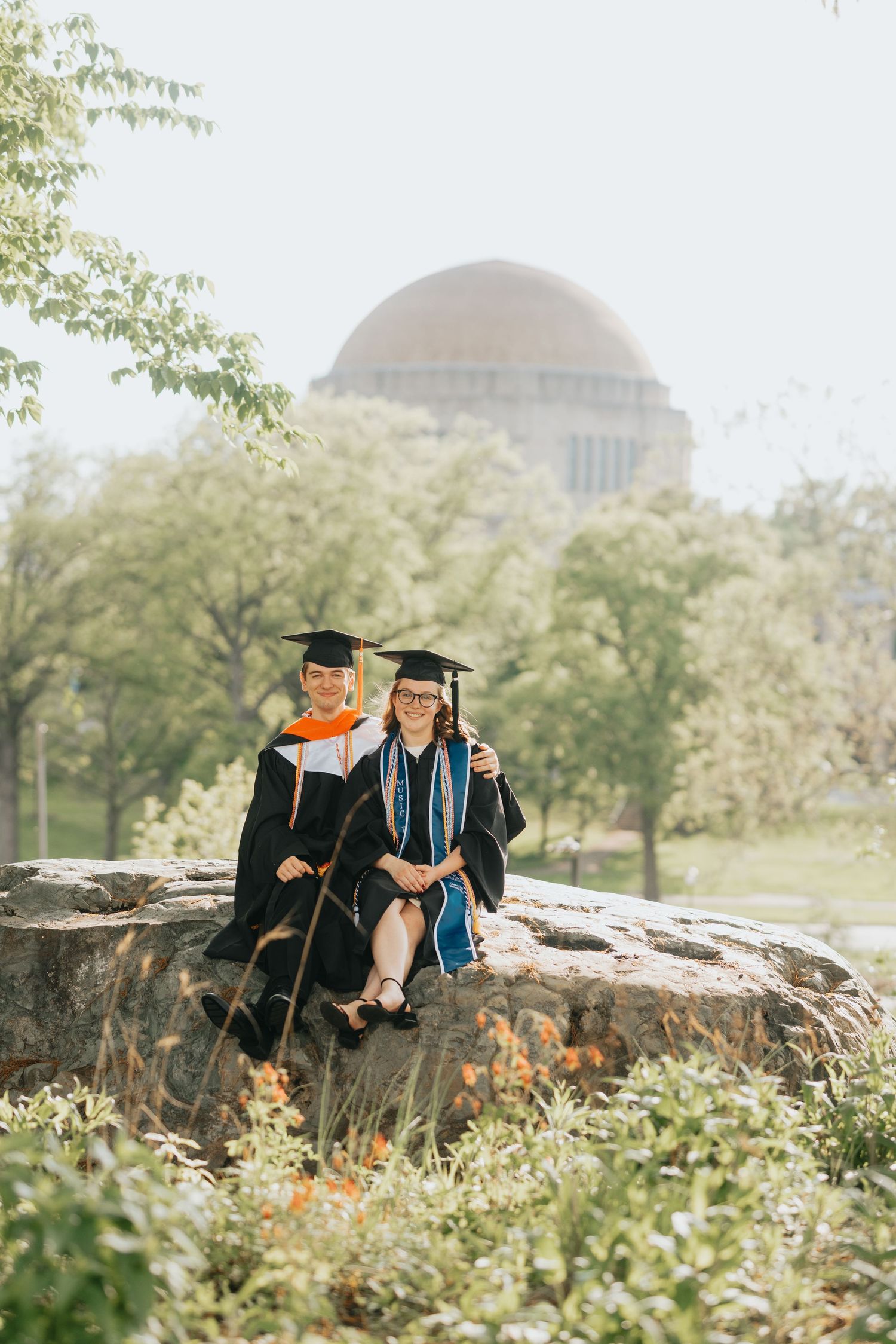 Two graduates in caps and gowns sitting on rocks with a university dome backdrop.