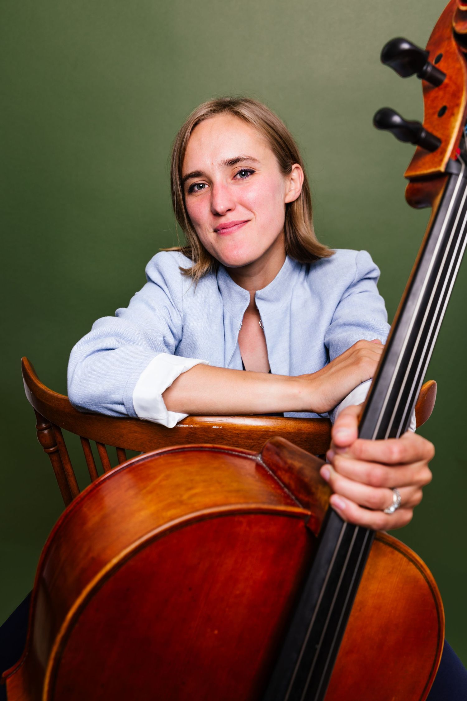 Smiling female cellist in a light blue blazer resting on her cello while seated against an olive green background.
