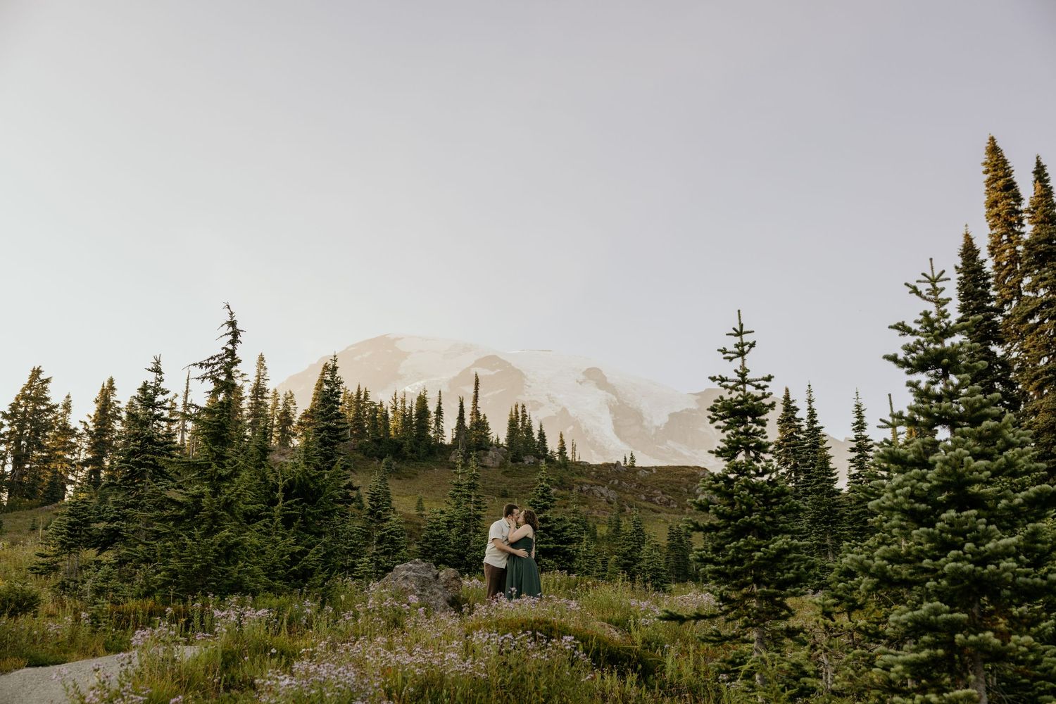 Couple embracing in a wildflower meadow surrounded by evergreen trees with Mount Rainier in the background.