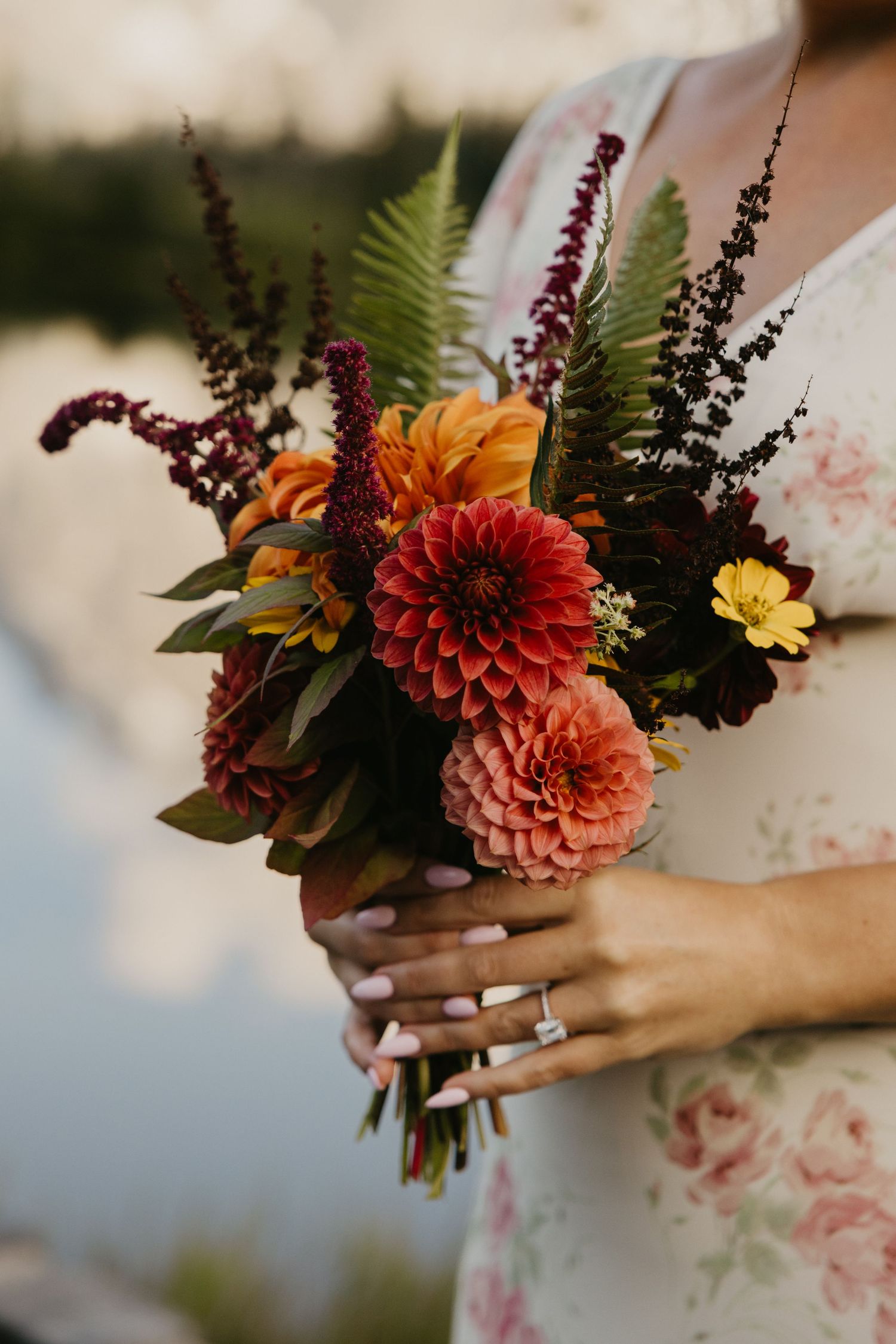 Bride in a floral dress holds an autumn bouquet of coral dahlias, burgundy amaranthus, and green ferns beside a calm lake.