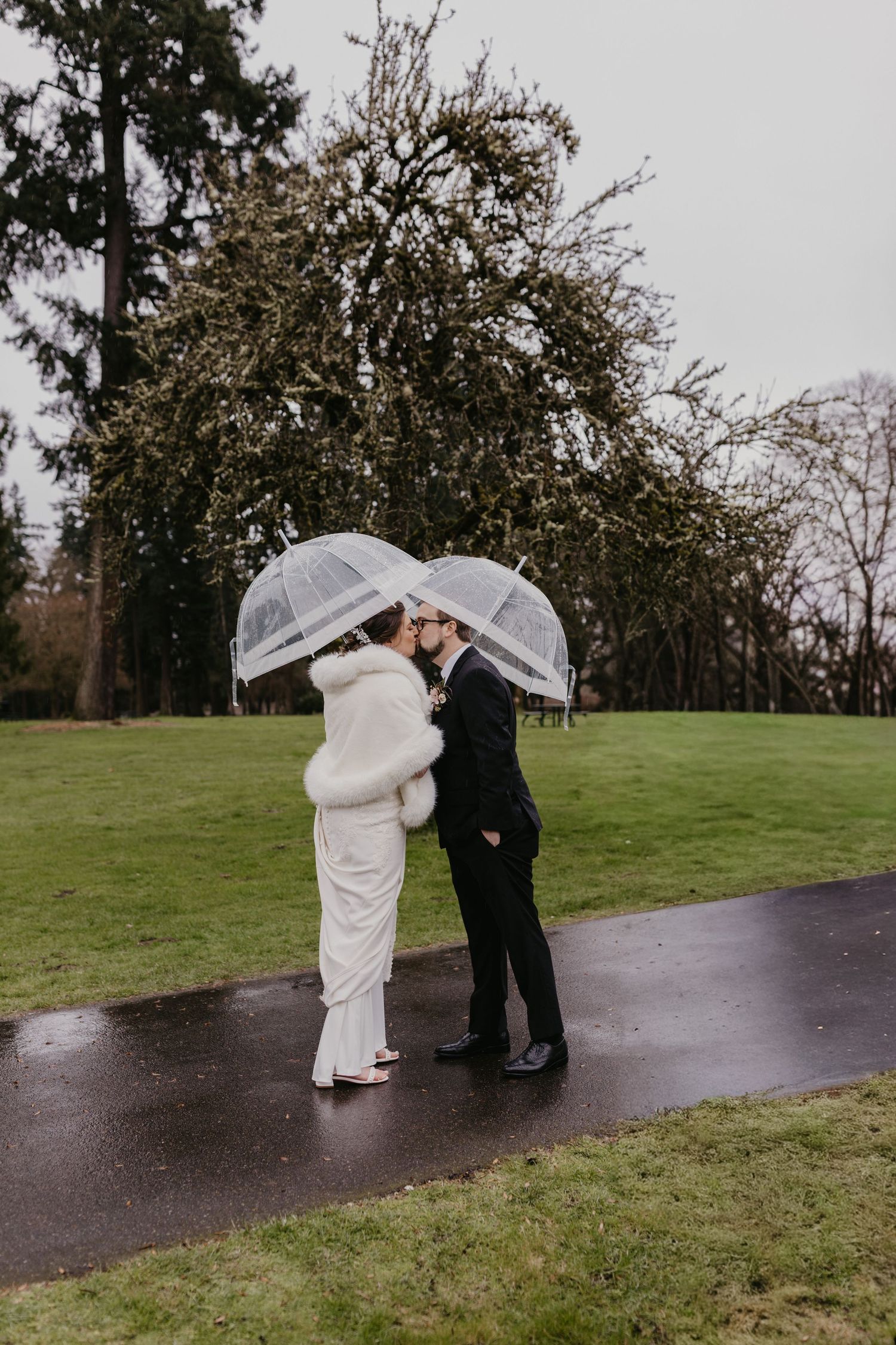 Bride and groom kissing under clear bubble umbrellas on a rainy wedding day in a lush green park.