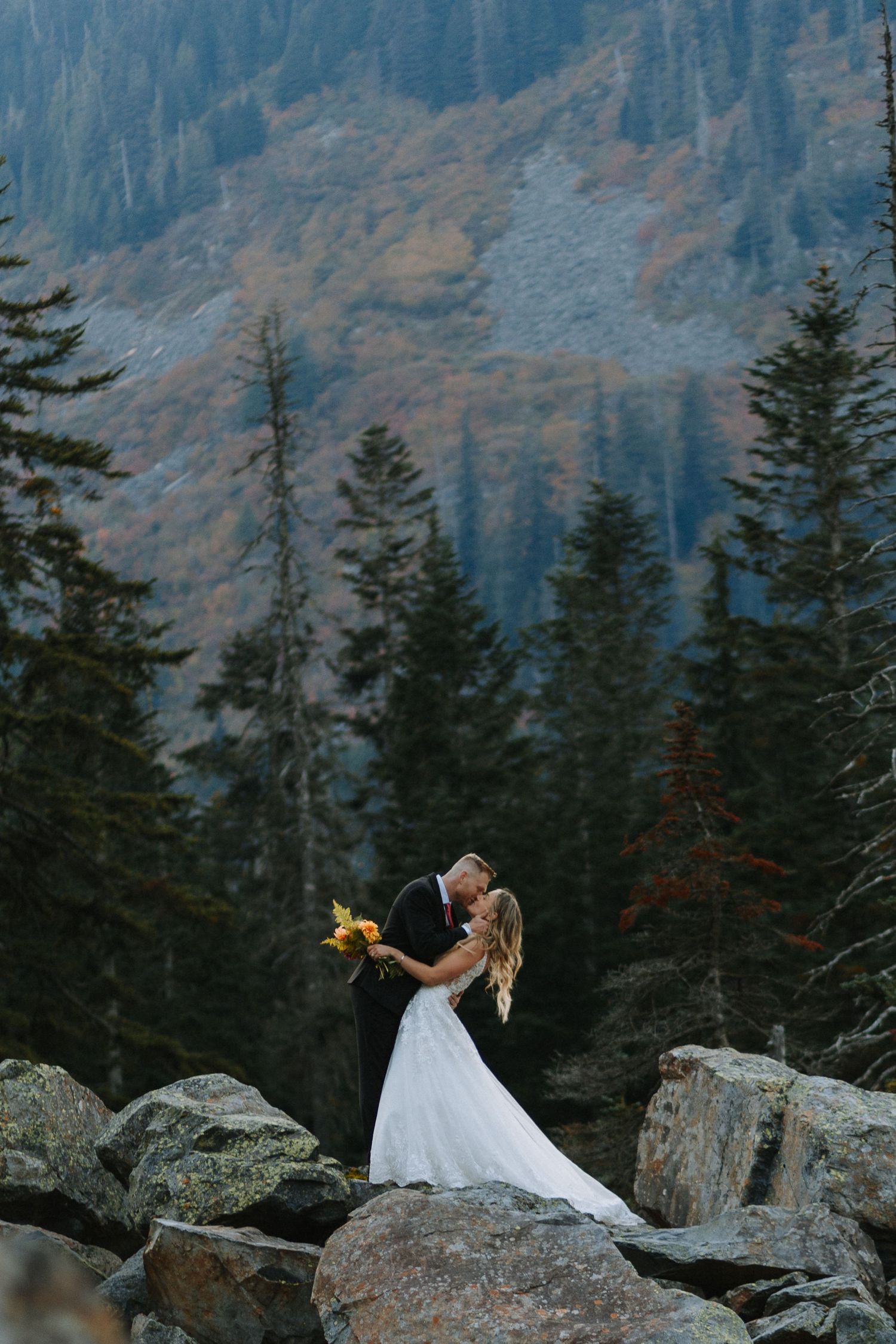 Bride and groom kissing on rocky mountain overlook surrounded by evergreen trees and autumn foliage near Seattle Washington