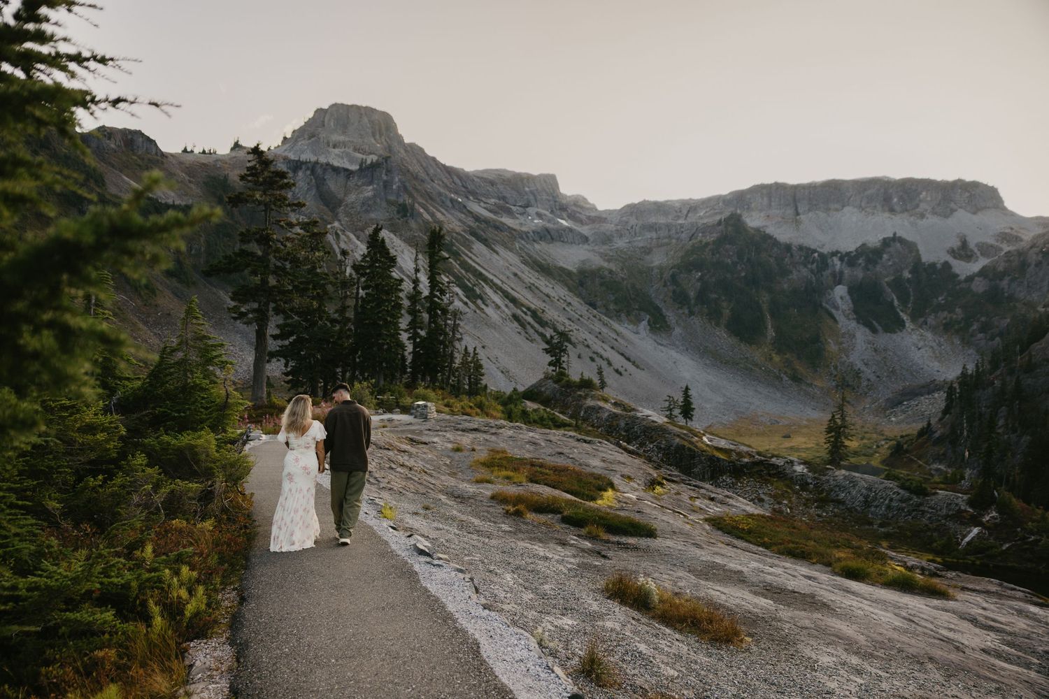 A bride and groom walk hand in hand along a mountain path surrounded by pine trees and rocky peaks at dusk.