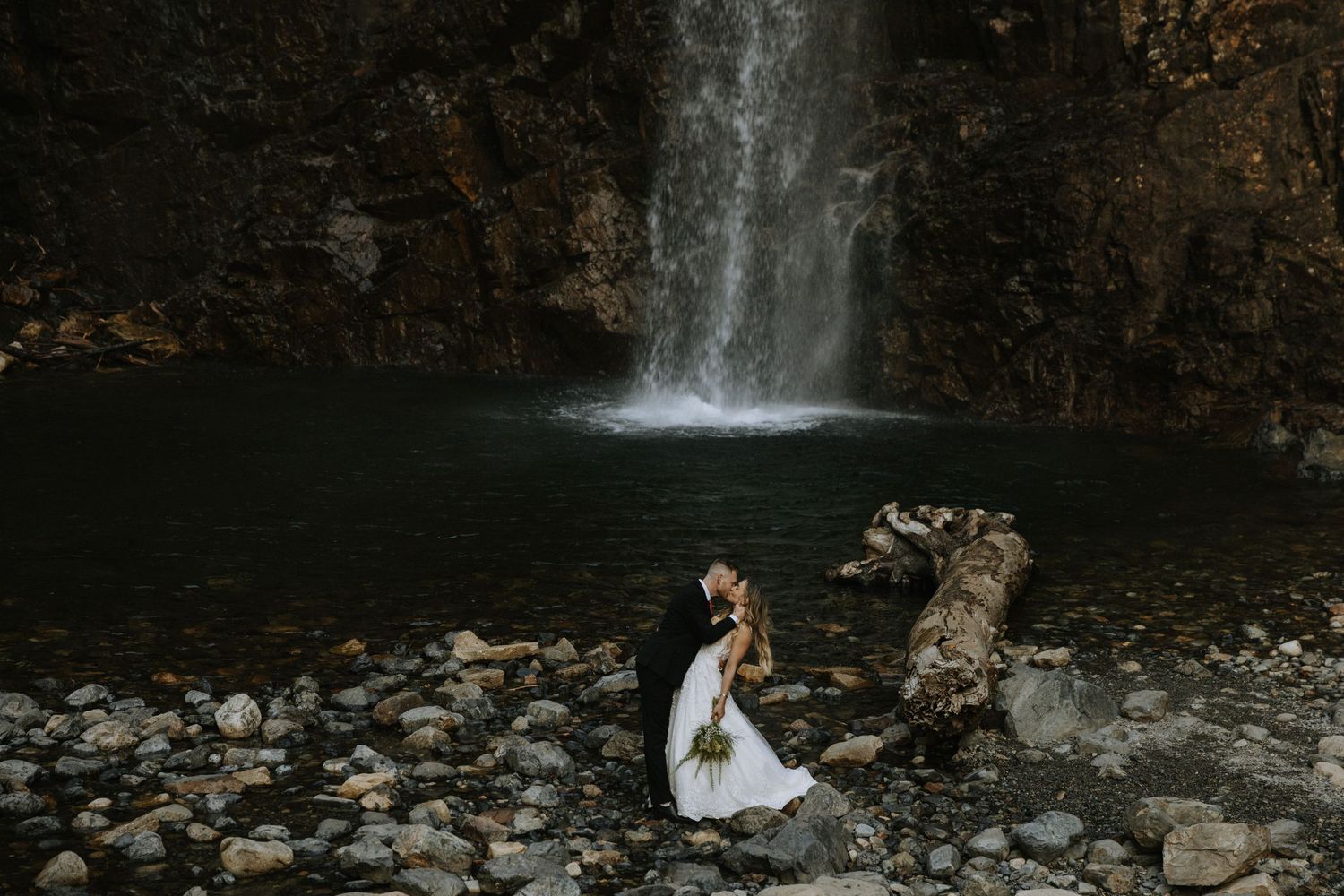 A bride and groom share a romantic moment on rocky shore before a stunning waterfall in a moody outdoor elopement.