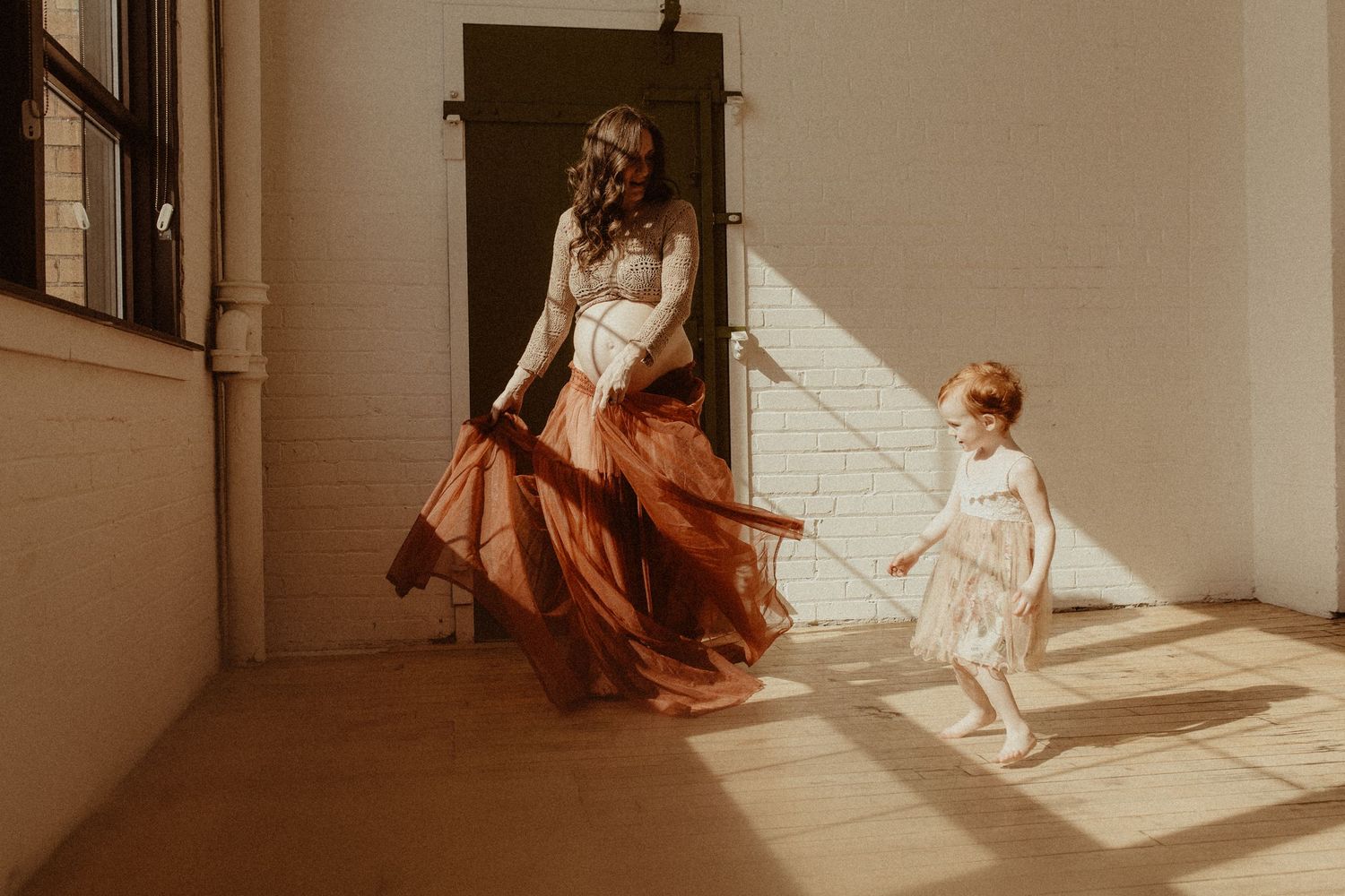 Pregnant mom in a long skirt dancing with her daughter in the harsh sunlight on old wooden floors by Bowmanville, ON family photographer