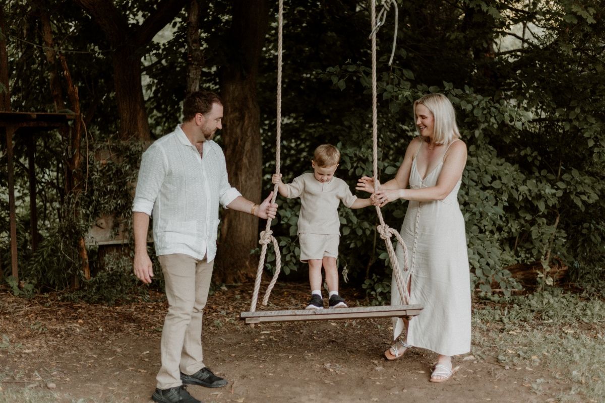 Family enjoying a session on a wooden swing, with parents smiling and standing close, by artistic family photographer Bowmanville, ON