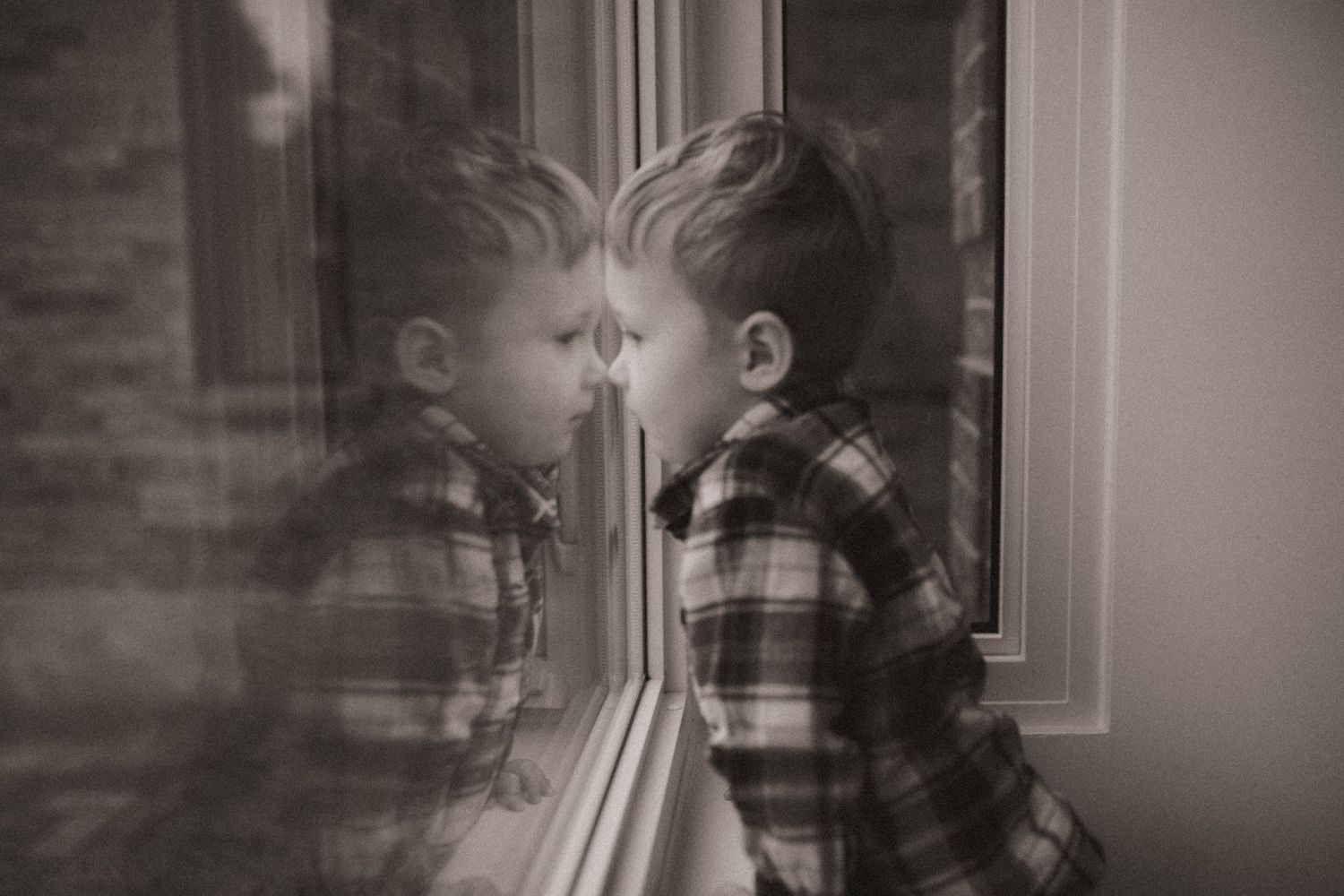 A boy in a plaid shirt sits by a window with his reflection visible in the glass, by artistic family photographer Bowmanville, ON.