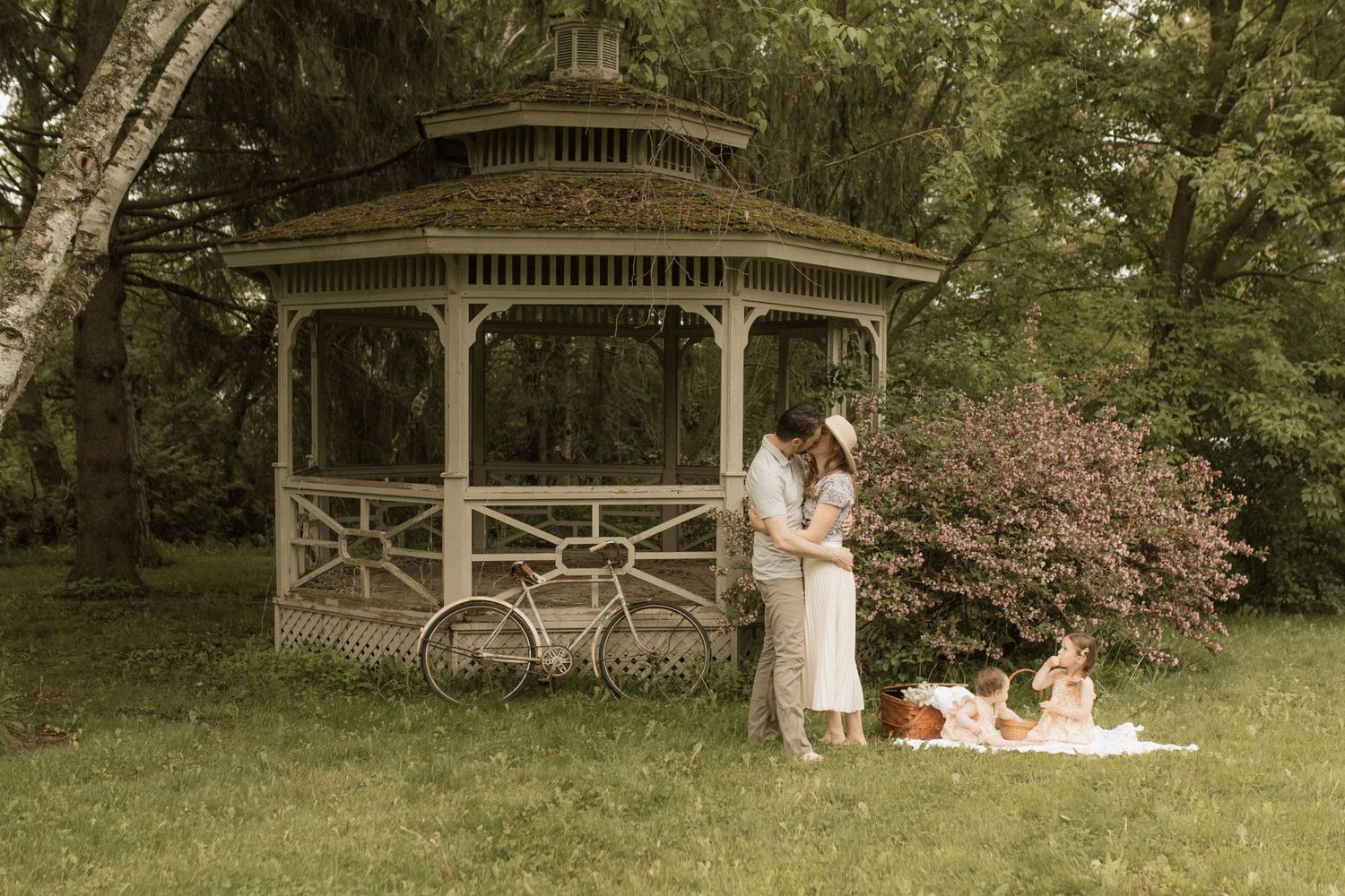 Young couple embracing in front of an antique gazebo while their young daughters are having a picnic by Bowmanville, ON family photographer.