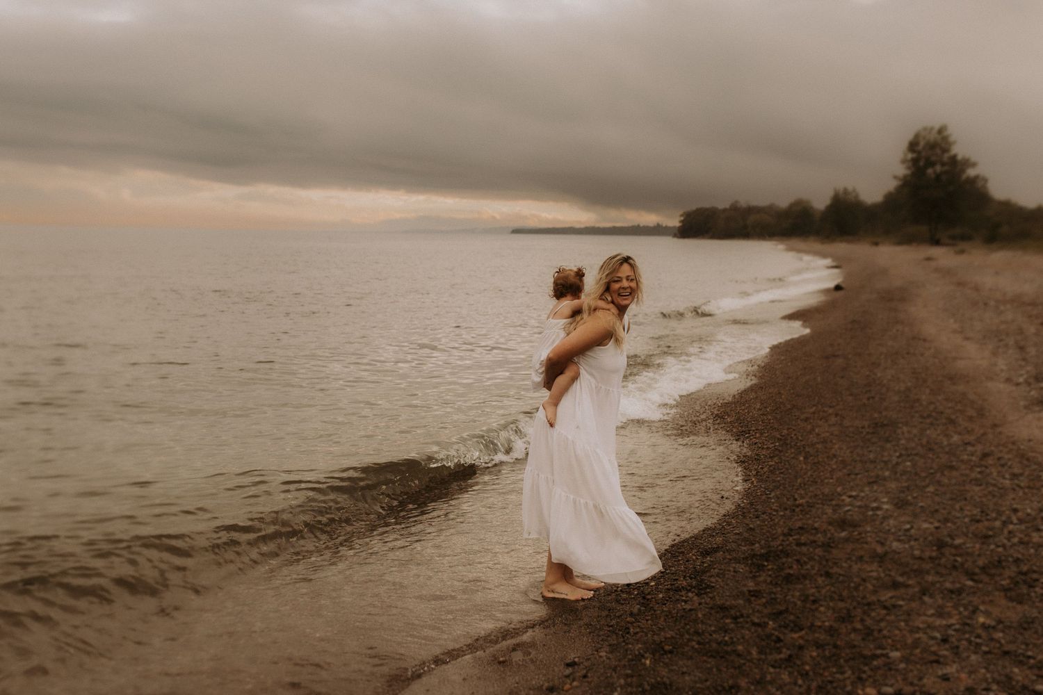 Mom carrying her toddler on her back, both wearing white dresses on a moody beach by Durham Region maternity photographer