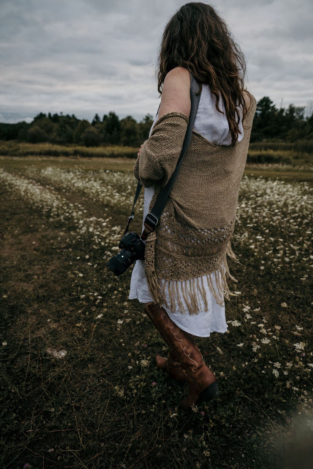 Your Bowmanville, ON family photographer in a white dress and brown shawl walking through a wildflower meadow at dusk.