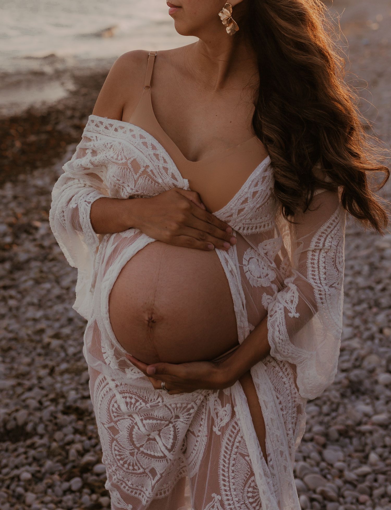 Expectant mother in a flowing dress, cradling her belly on the sand as waves roll in.