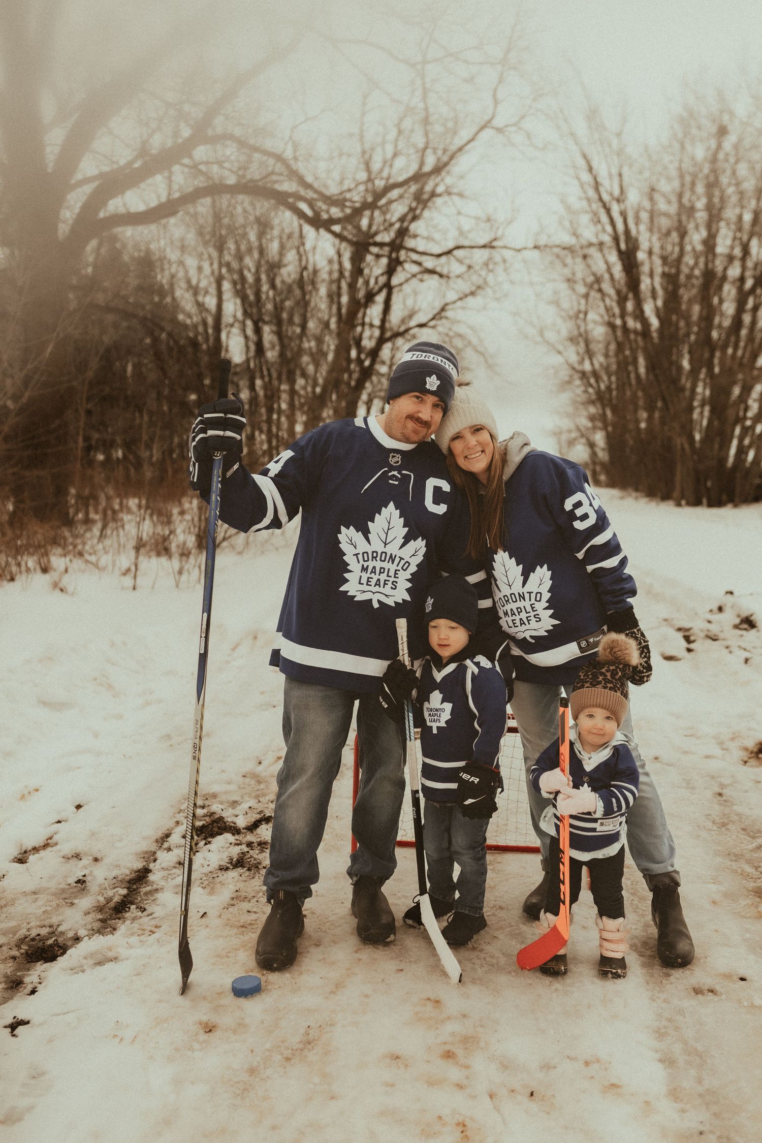 Hockey family in winter posing with hockey sticks in their Maple Leaf jerseys by Newcastle, ON family photographer