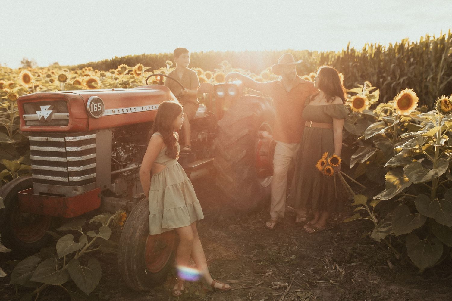Golden hour photo of a young family gathered around an antique tractor in a sunflower field by Durham Region Family Photographer