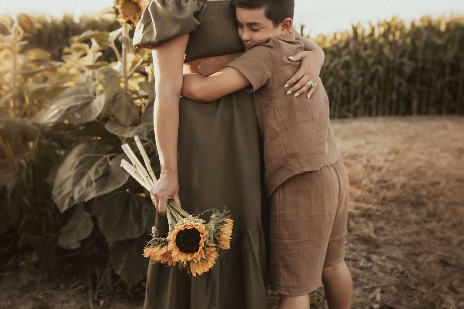 A young boy giving his mom a tight hug in a sunflower field as his mom holds a bouquet of sunflowers by Durham Region family photographer