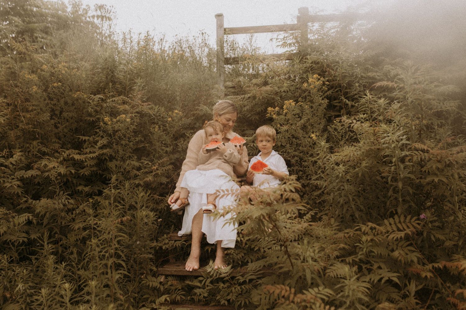 A mom with her two children enjoying a slice of watermelon on the wooden, overgrown steps near the beach by Cobourg family photographer