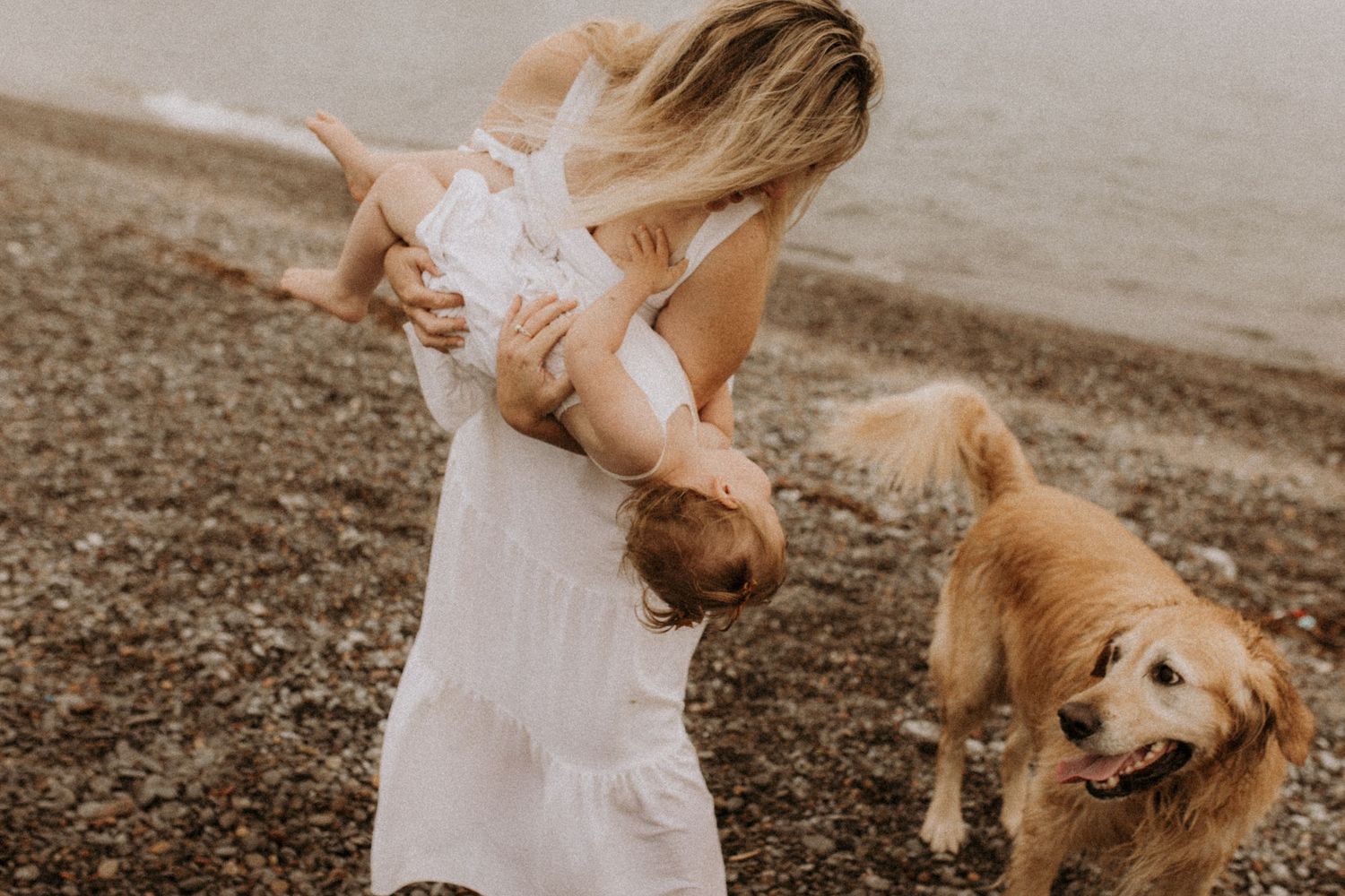 A playful moment between a mom, daughter and dog on an overcast summer beach by Cobourg family photographer