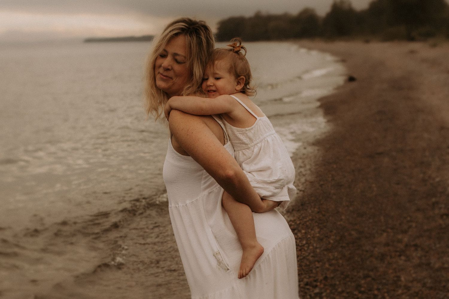 Mom holding daughter on her back and laughing playfully on a moody beach by Cobourg family photographer