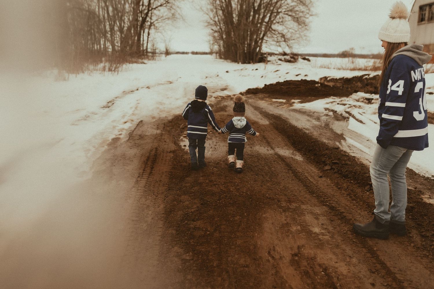 Photo of two young kids playing in the mud in their hockey jerseys as their mom looks on by Newcastle, ON family photographer