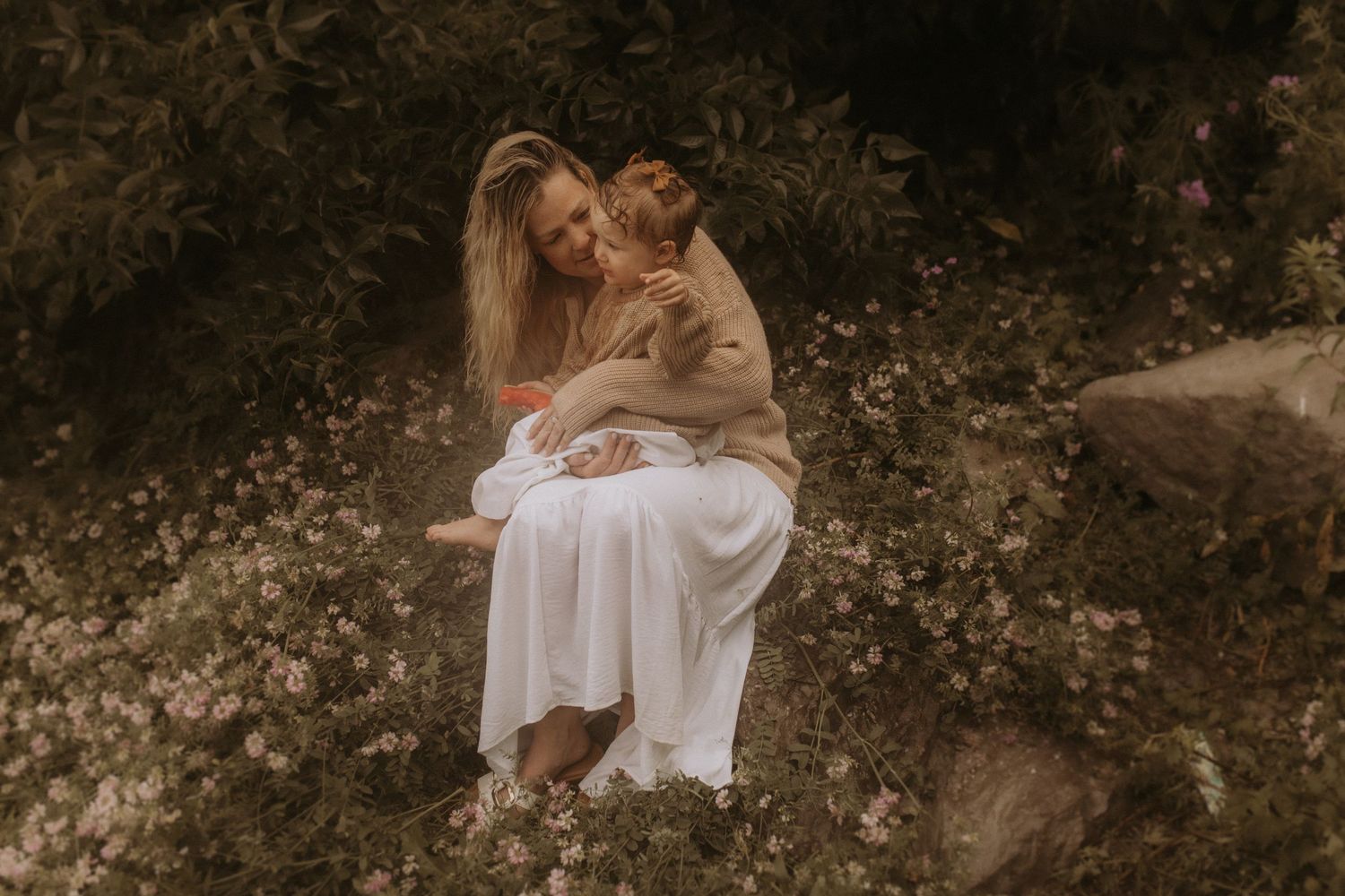 A mom cuddling her son on the large rocks surrounded by flowers near a beach by Cobourg family photographer