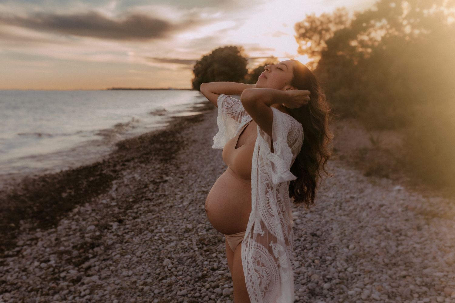 Sunset casting a warm glow on a pregnant woman’s silhouette, standing barefoot on the beach.