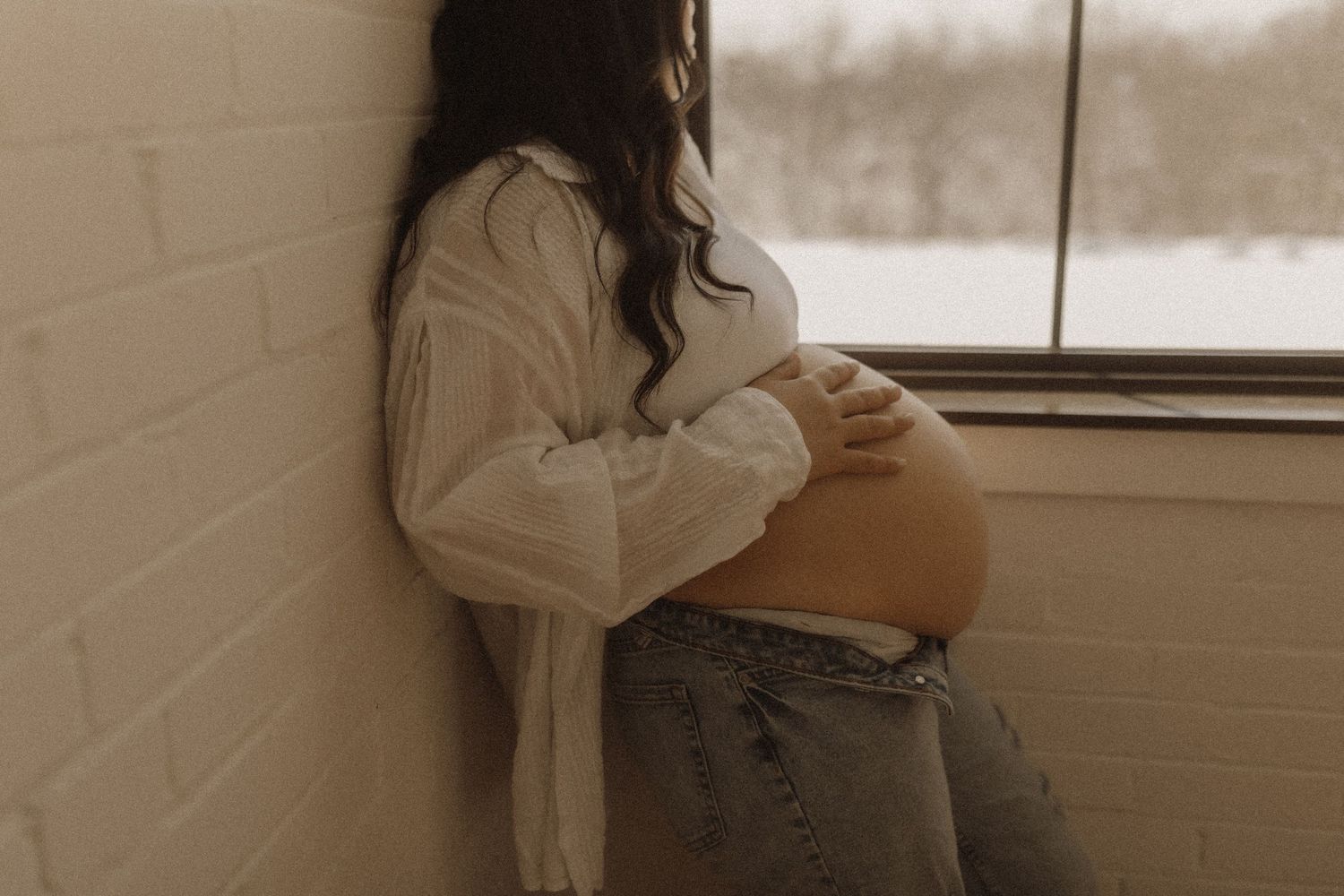 Pregnant woman gazing out a wintery window cradling her baby bump gently by Durham Region maternity photographer