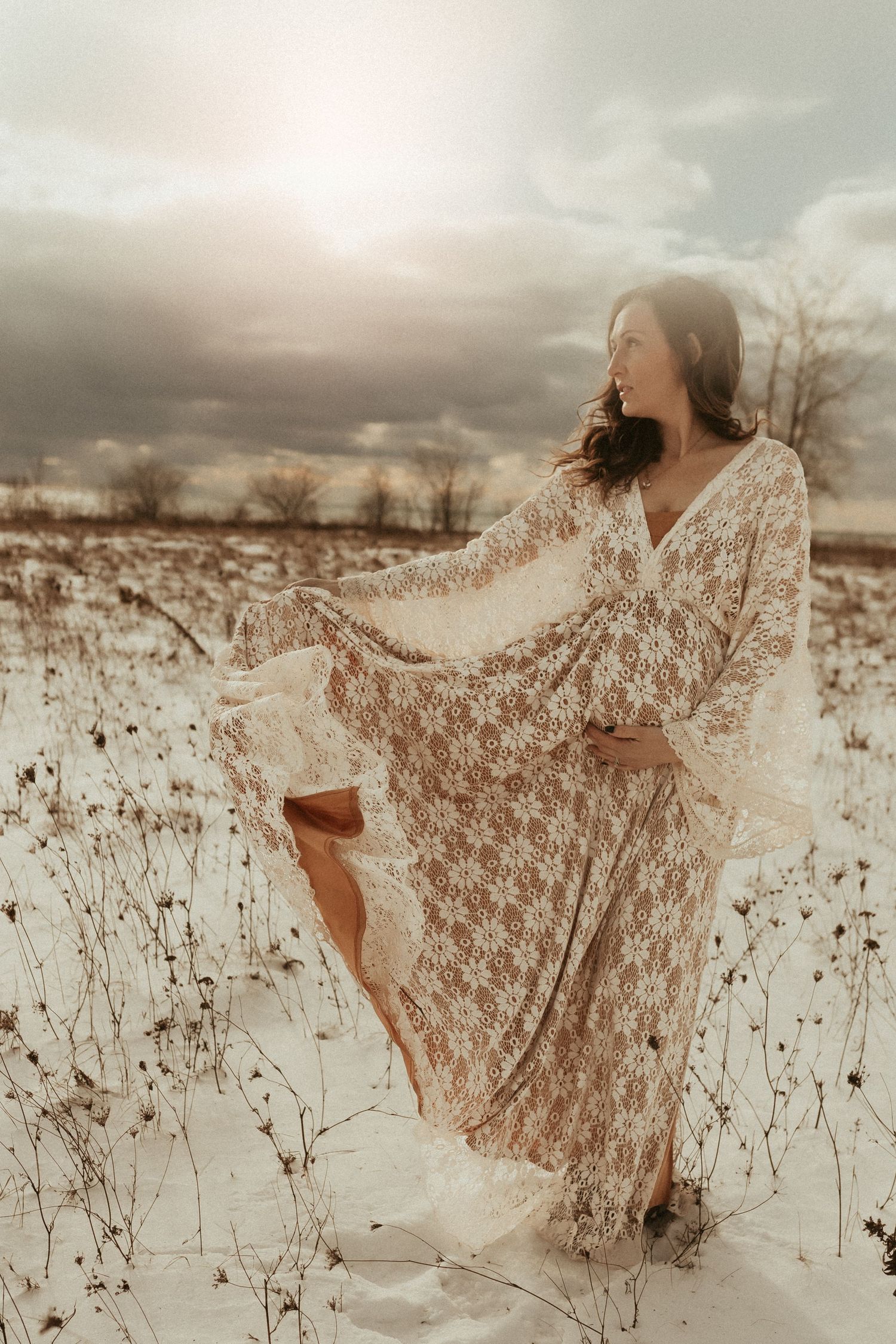 A figure in a flowing white lace dress dances gracefully through a snowy winter field at sunset.