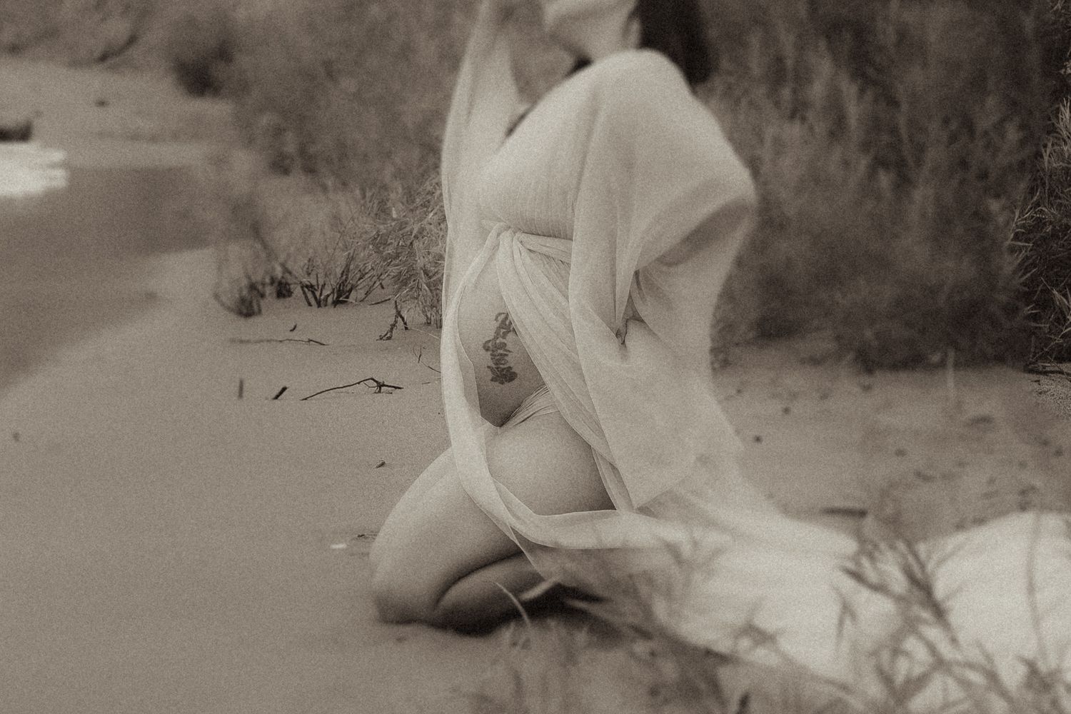 Mom-to-be kneeling on a beach in a long, wispy dress by Durham Region maternity photographer