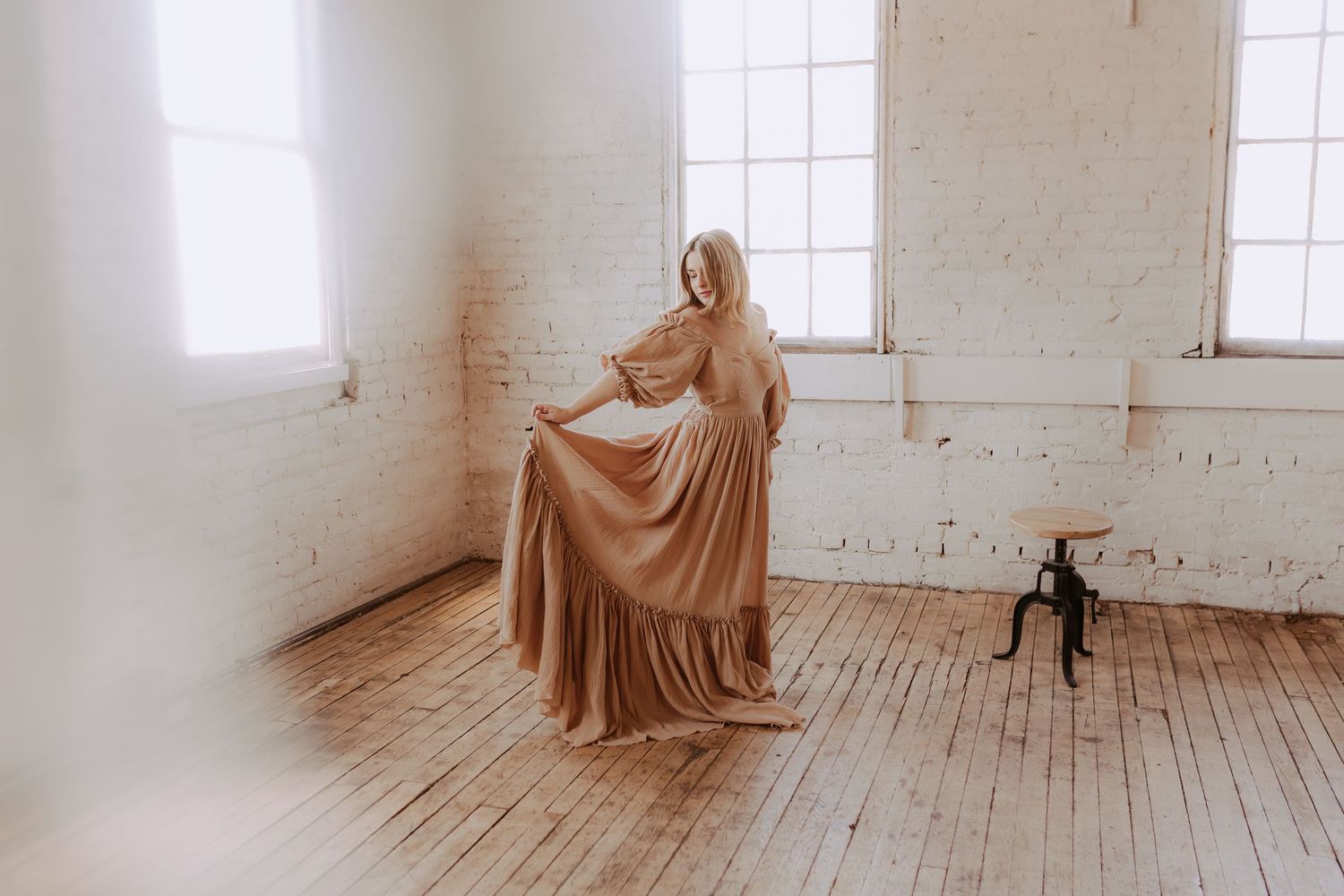 Flowing brown dress creates dramatic silhouette against white walls and wooden floor in minimalist studio space.