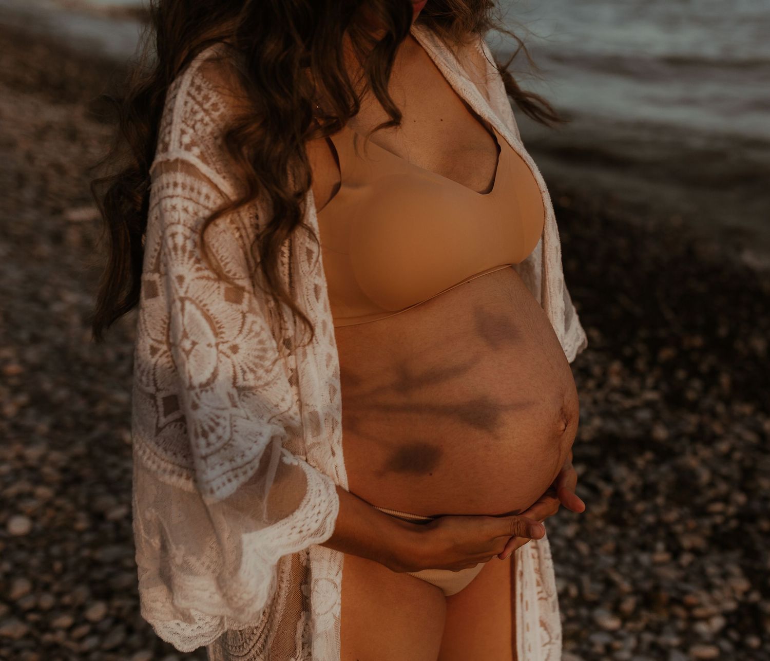 Pregnant woman standing on the beach at sunset, gently holding her baby bump.