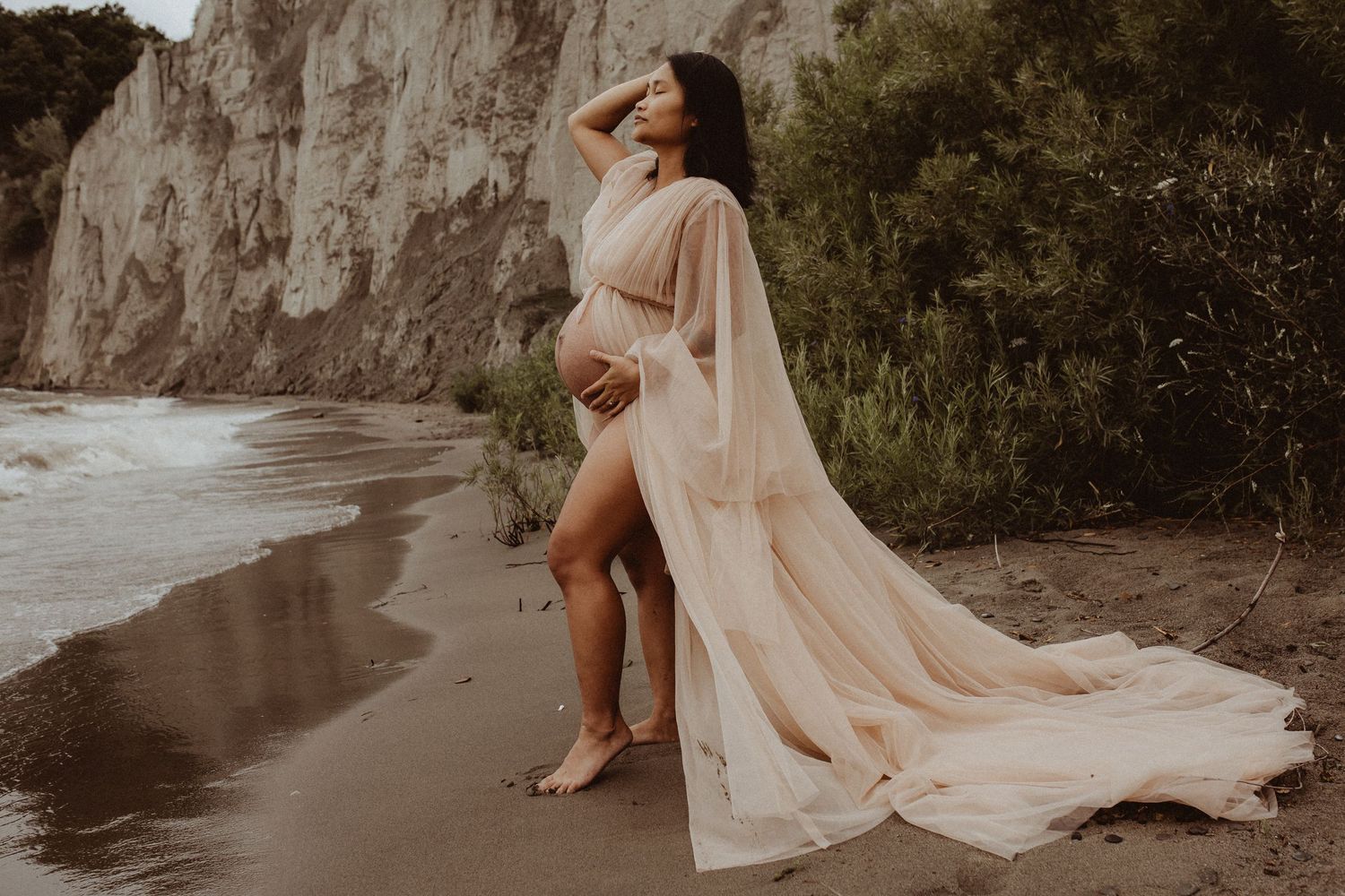 A dramatic photo of a pregnant mom in a flowing gown on a beach with cliffs behind her during a Bowmanville maternity photography session.