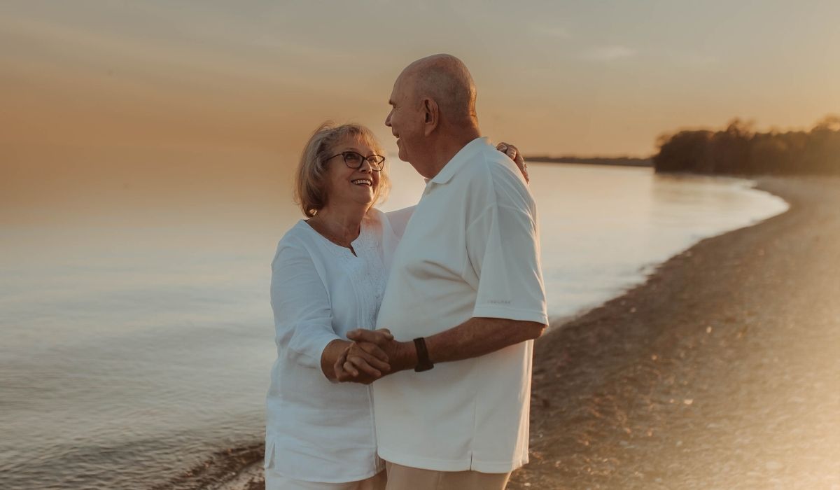 Elderly couple dancing together on the beach at sunset, holding each other closely as the golden light reflects off the waves.