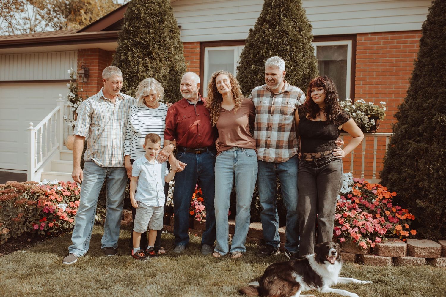 A multi-generational family stands together in front of a brick house with blooming red flowers in the garden.