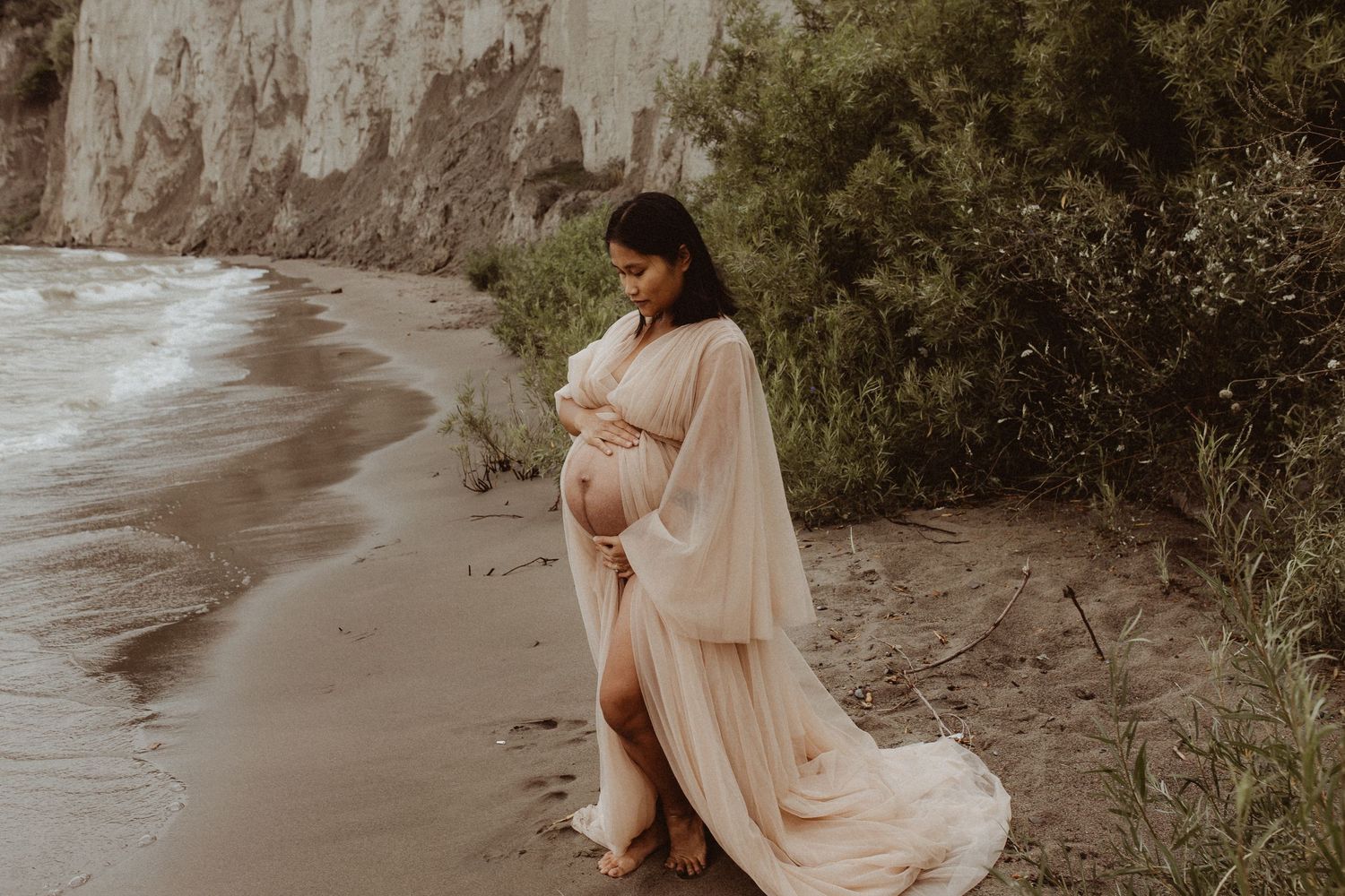 A mom looking lovingly down at her pregnant belly on the beach during a Bowmanville maternity photography session.