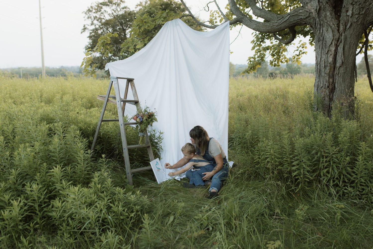 A wide-shot image of a mom and son painting together in a field in front of a white sheet with an old ladder by Courtice family photographer