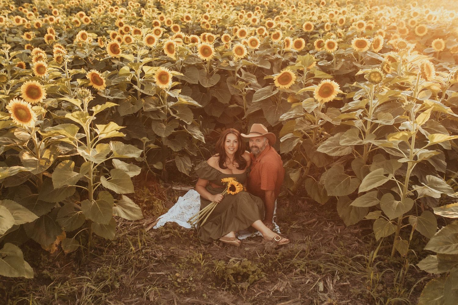 A husband and wife smile at the camera in a field of sunflowers by Durham Region family photographer