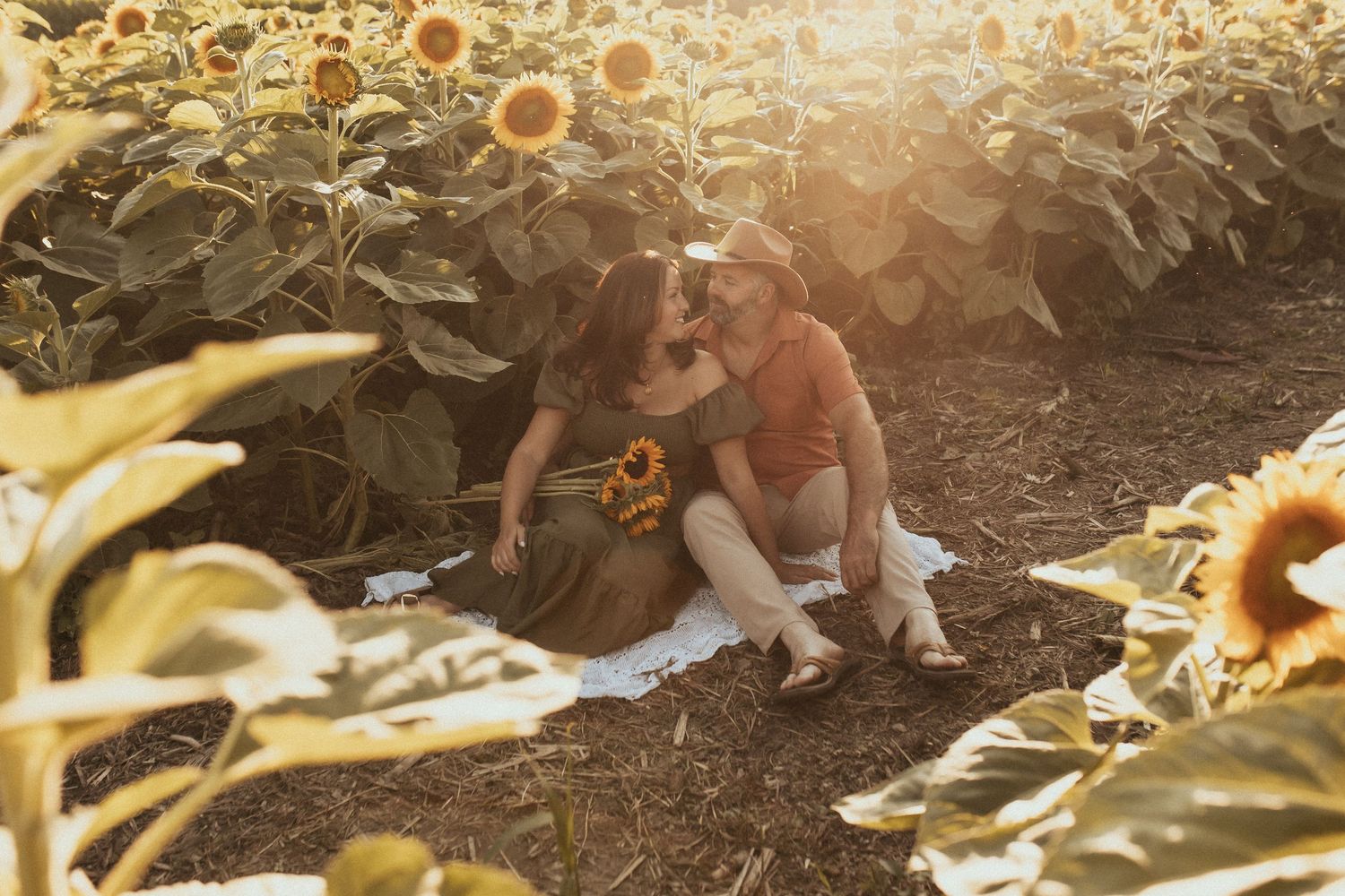 A photo of a couple sitting on a blanket in a sunflower field during sunset by Durham Region family photographer