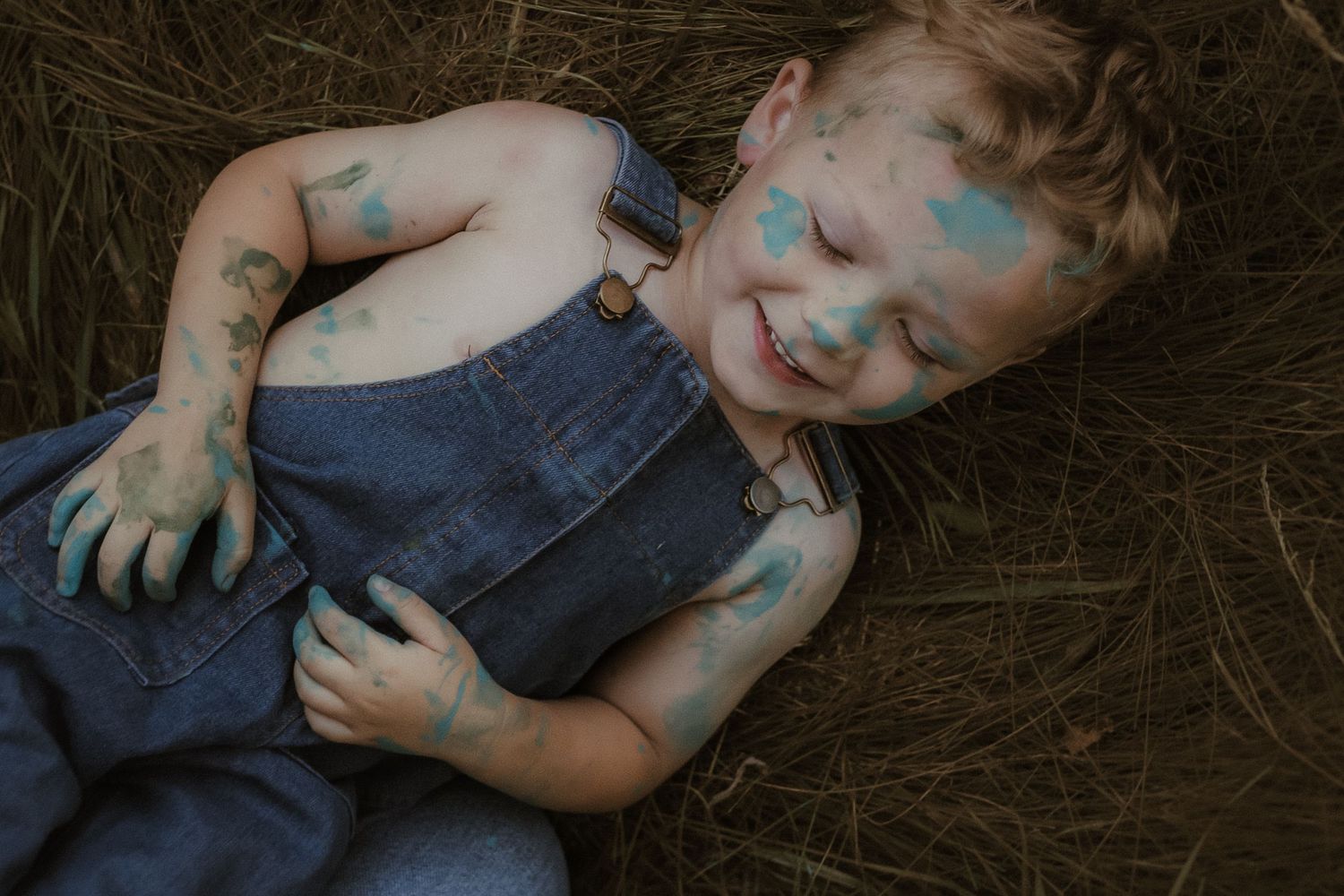 A young boy is lying in the grass smiling to himself with a face covered in paint by Courtice family photographer