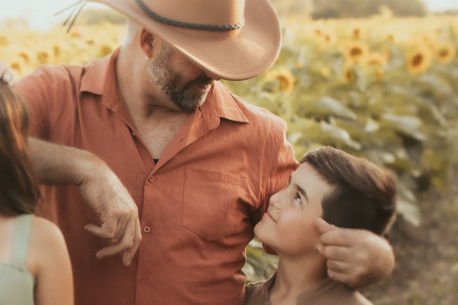 A man in a cowboy hat looks down affectionately at his son in a field of sunflowers by Durham Region family photographer