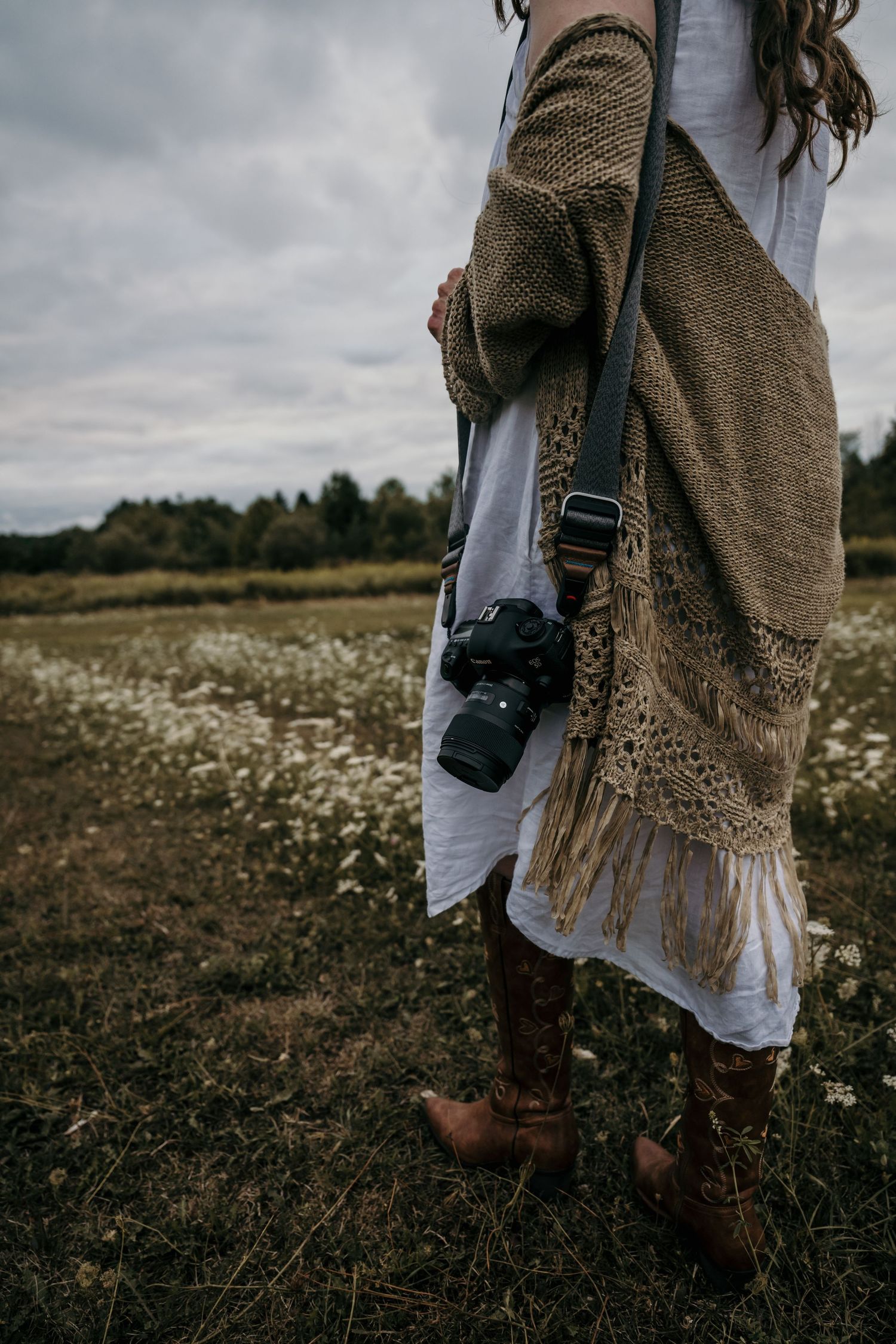 Person in white dress and brown shawl photographing in a daisy-filled meadow.