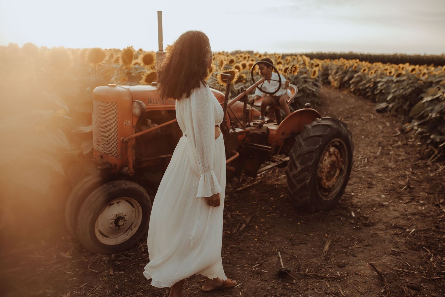 A mother in a long white dress watches on as her son plays on an tractor in a sunflower field by Durham Region maternity photographer