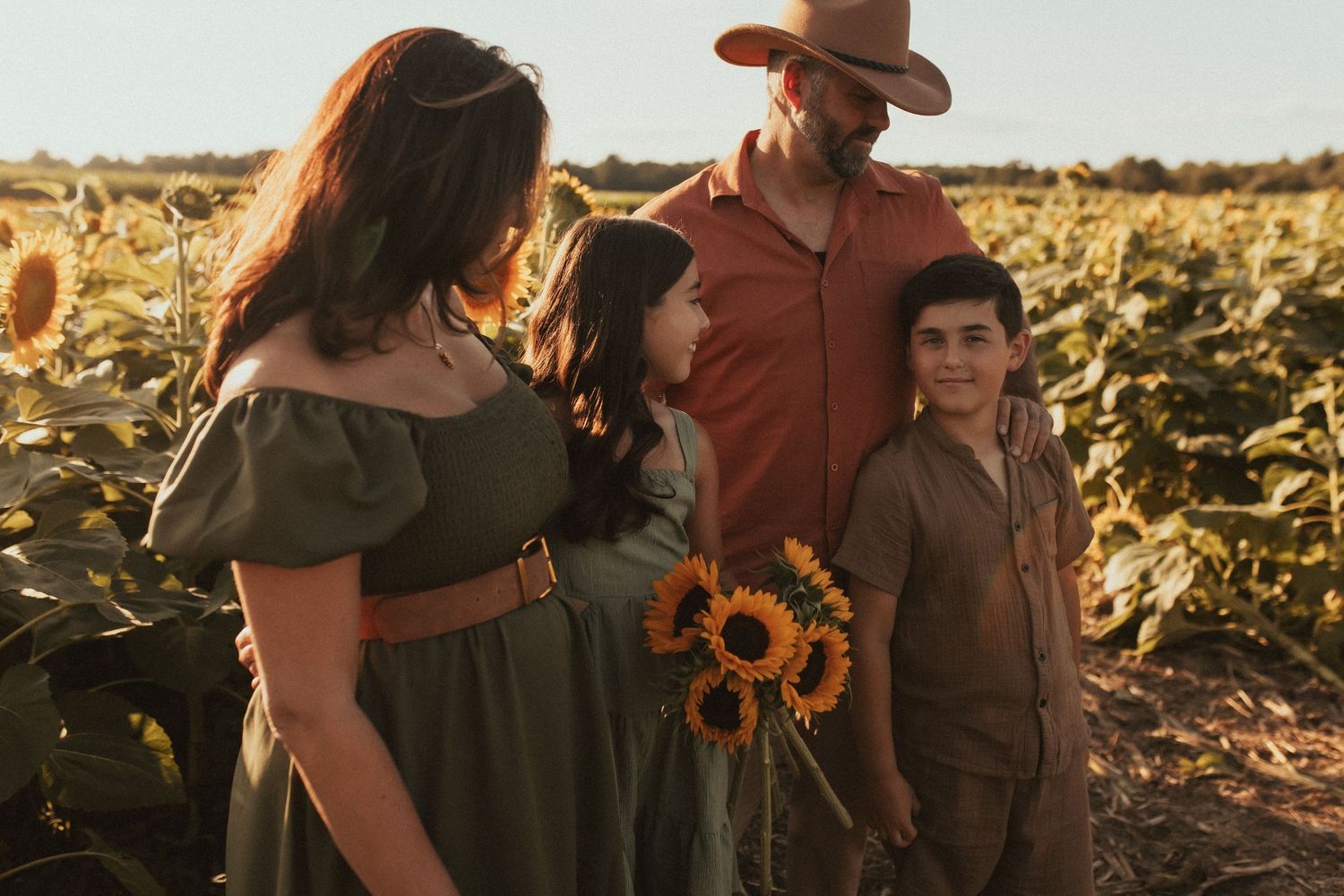 A faraway shot of a family standing close together in a sunflower field drenched in golden light by Durham Region family photographer