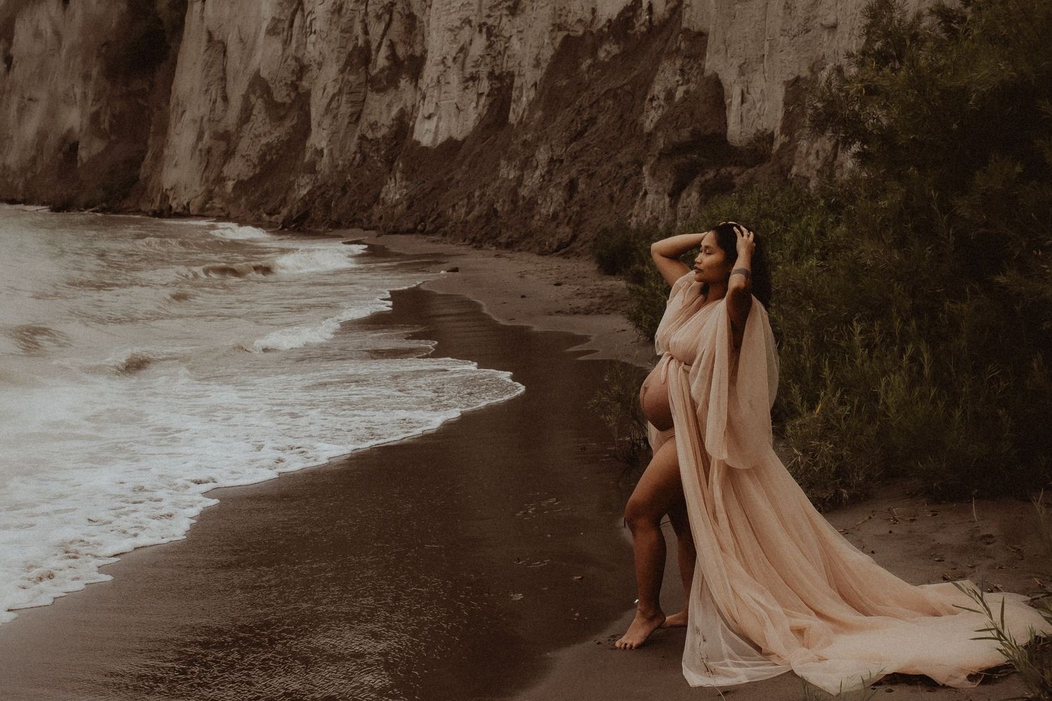 Expectant mother in a long, flowy gown standing on a beach at the Scarborough Bluffs by Bowmanville maternity photographer