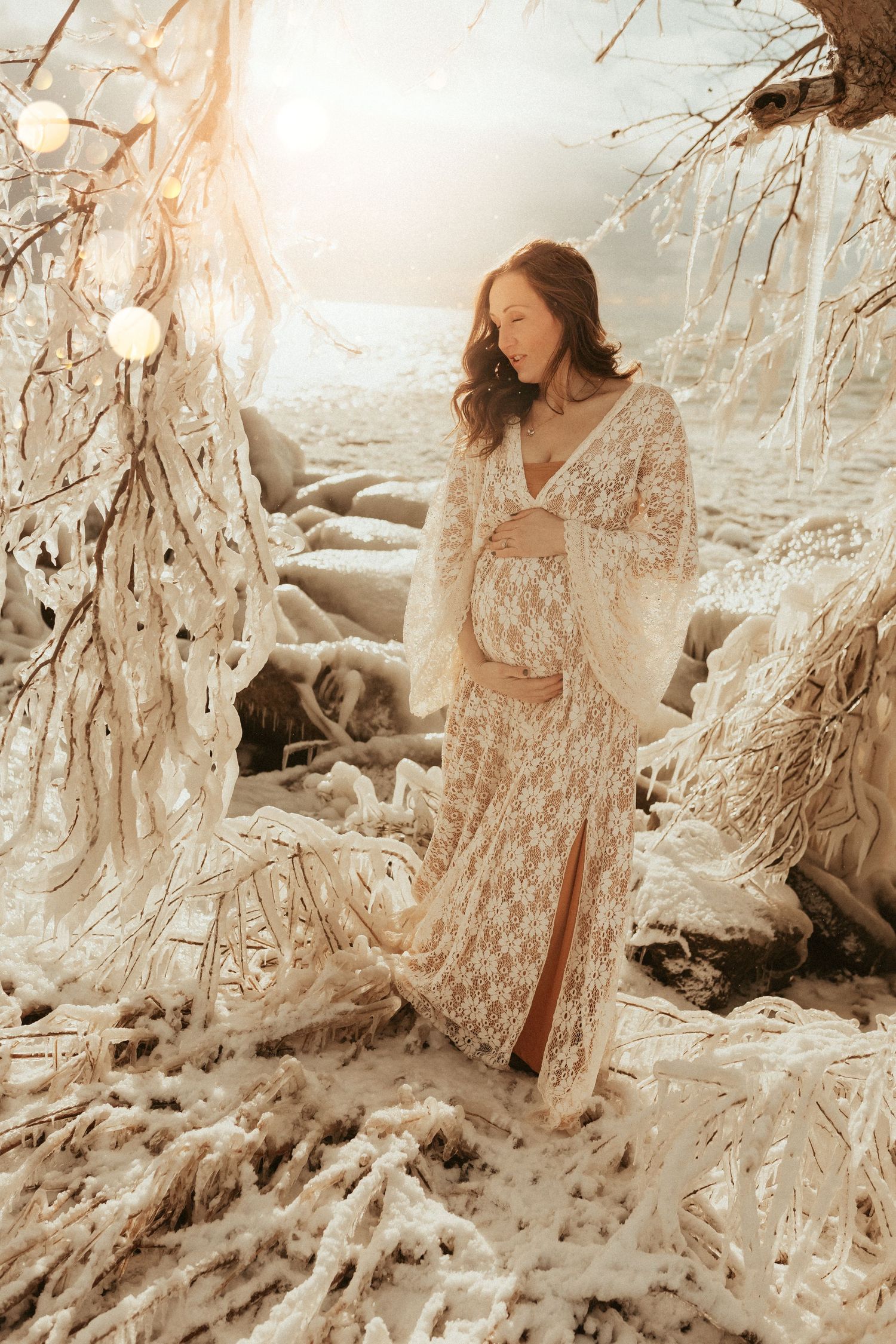 Ethereal maternity photoshoot featuring  a delicate white lace dress in a dreamy winter wonderland setting with icy branches.