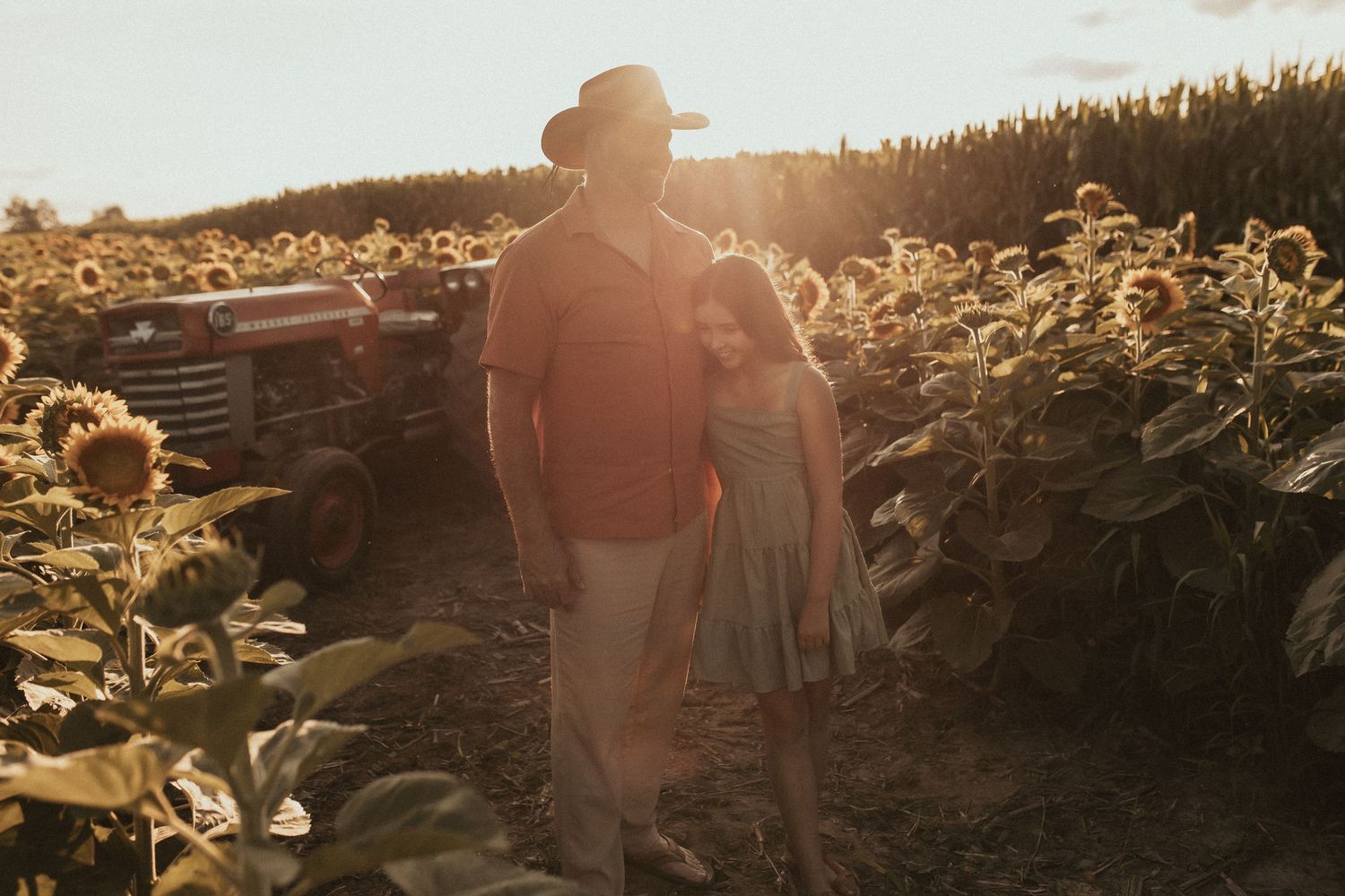 A father and daughter quietly hug while standing near the antique tractor in a sunflower field by Durham Region family photographer