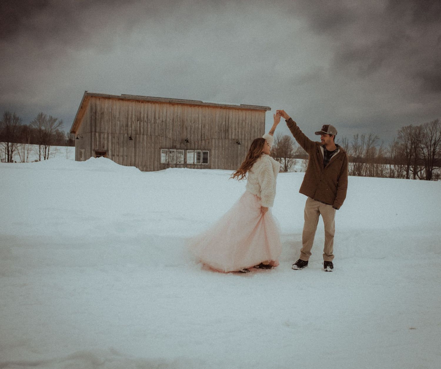 A couple dances together in a snowy winter landscape with rustic barns in the background, by artistic family photographer Bowmanville, ON.