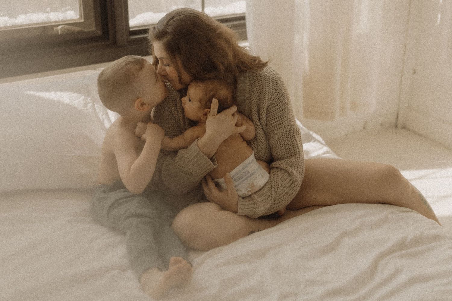 Mom kissing her little boy as she also holds her newborn in her arms on a bed taken by artistic family photographer Bowmanville, ON.