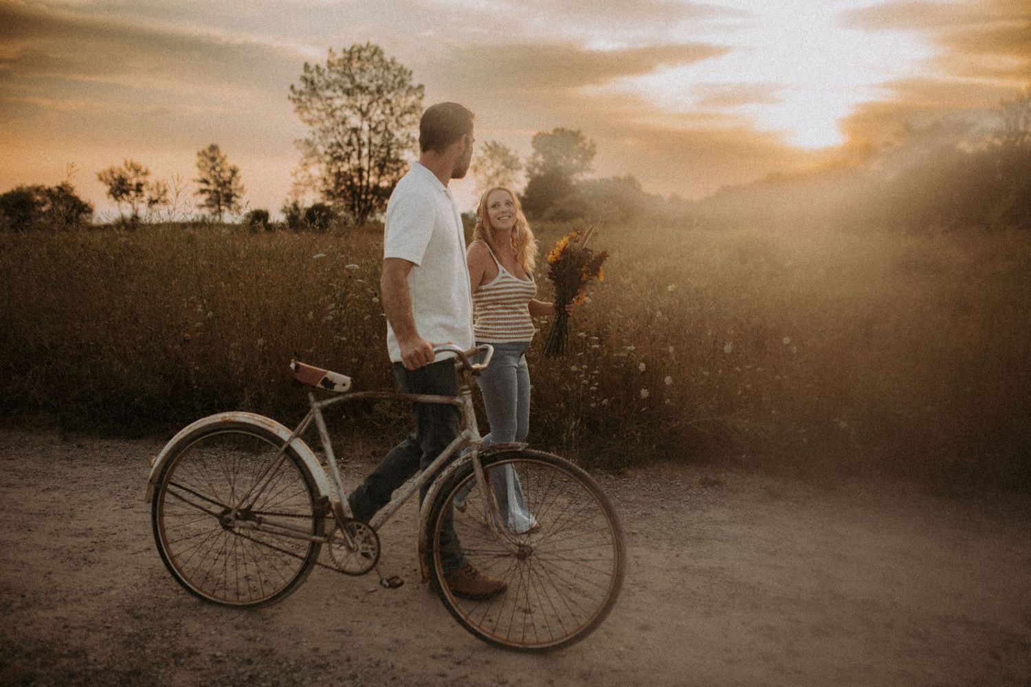 Boho couple walking a vintage bike in a sunlit field during golden hour by family photographer Bowmanville, ON