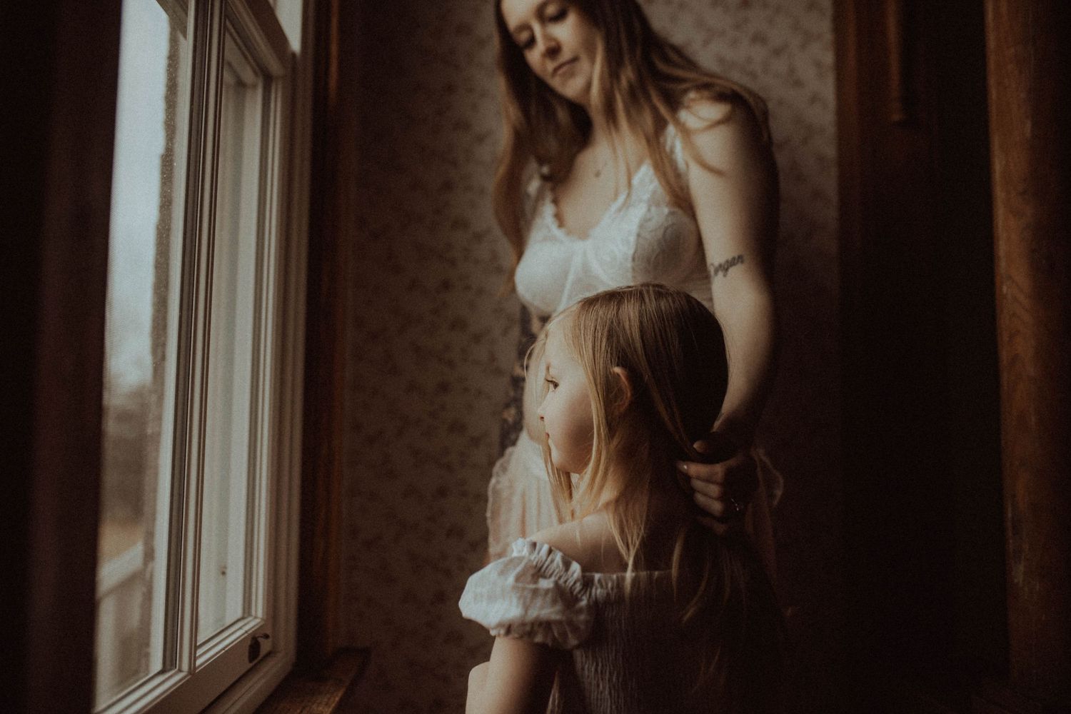 Image of an expectant mom gently touching her daughter's hair in front of a window by Bowmanville, ON family photographer.