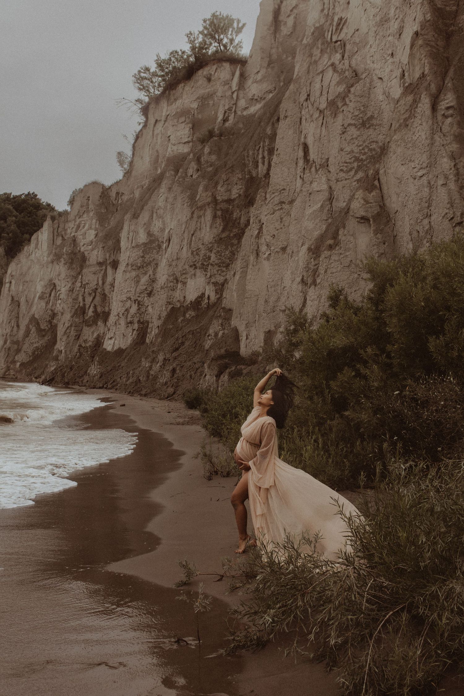 Pregnant mom playing with her hair on a beach in front of a cliff by Durham Region maternity photographer