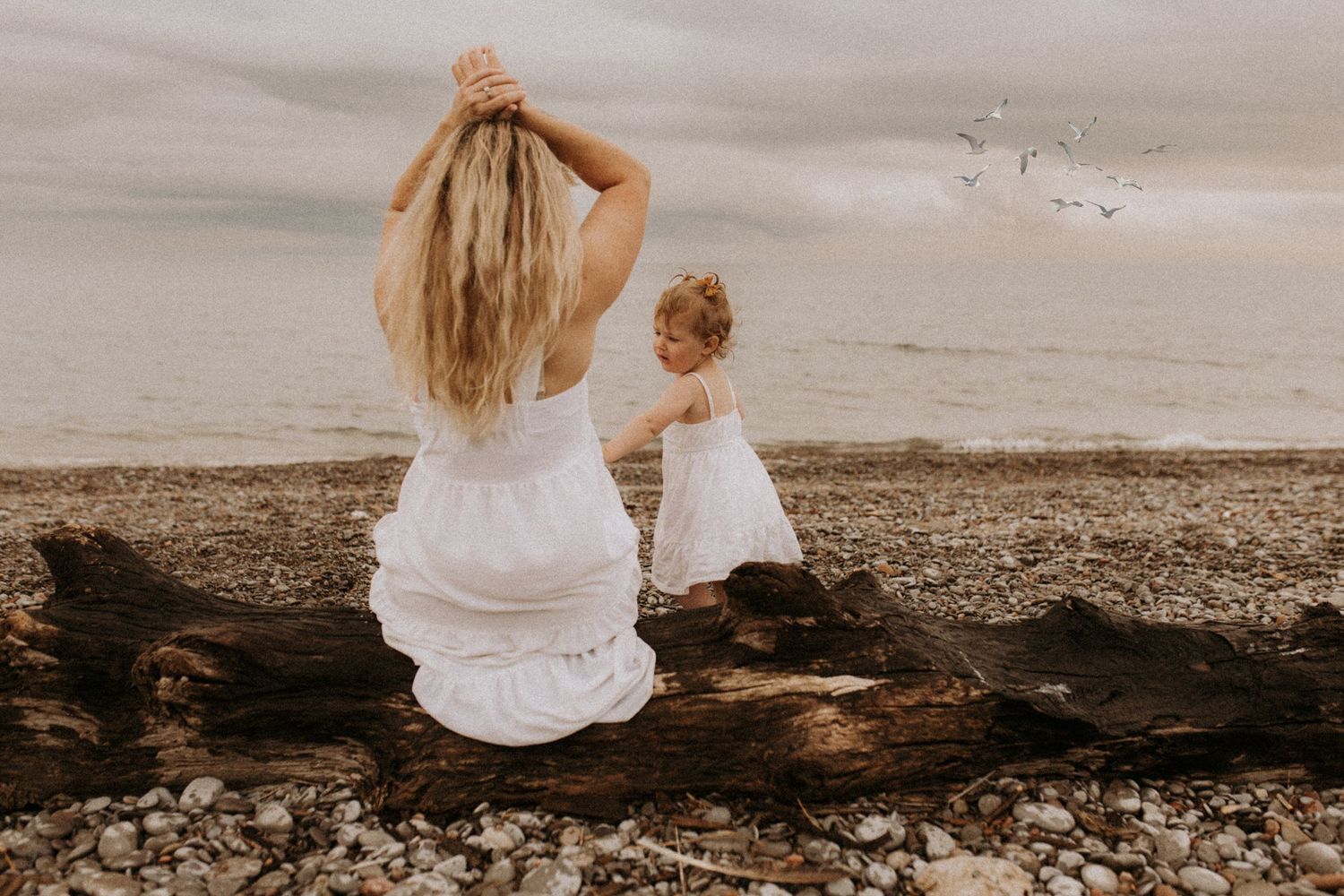 Mom sitting on a log on the beach and watching her daughter enjoy the moment by by Cobourg family photographer